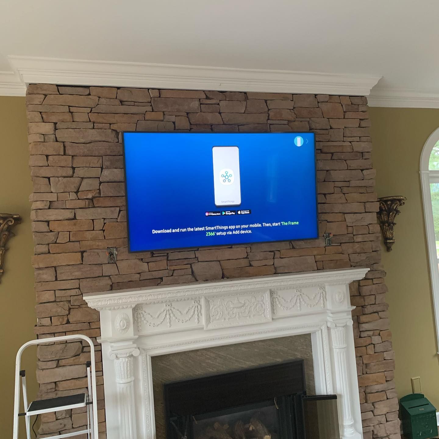 TV mounted on a stone fireplace above a white mantel. A stepladder sits nearby.