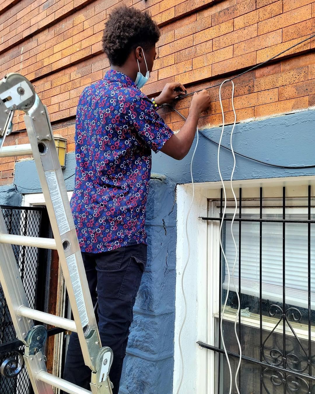 Person on ladder attaching wires to a brick building exterior. Wears a face mask and button-down shirt.
