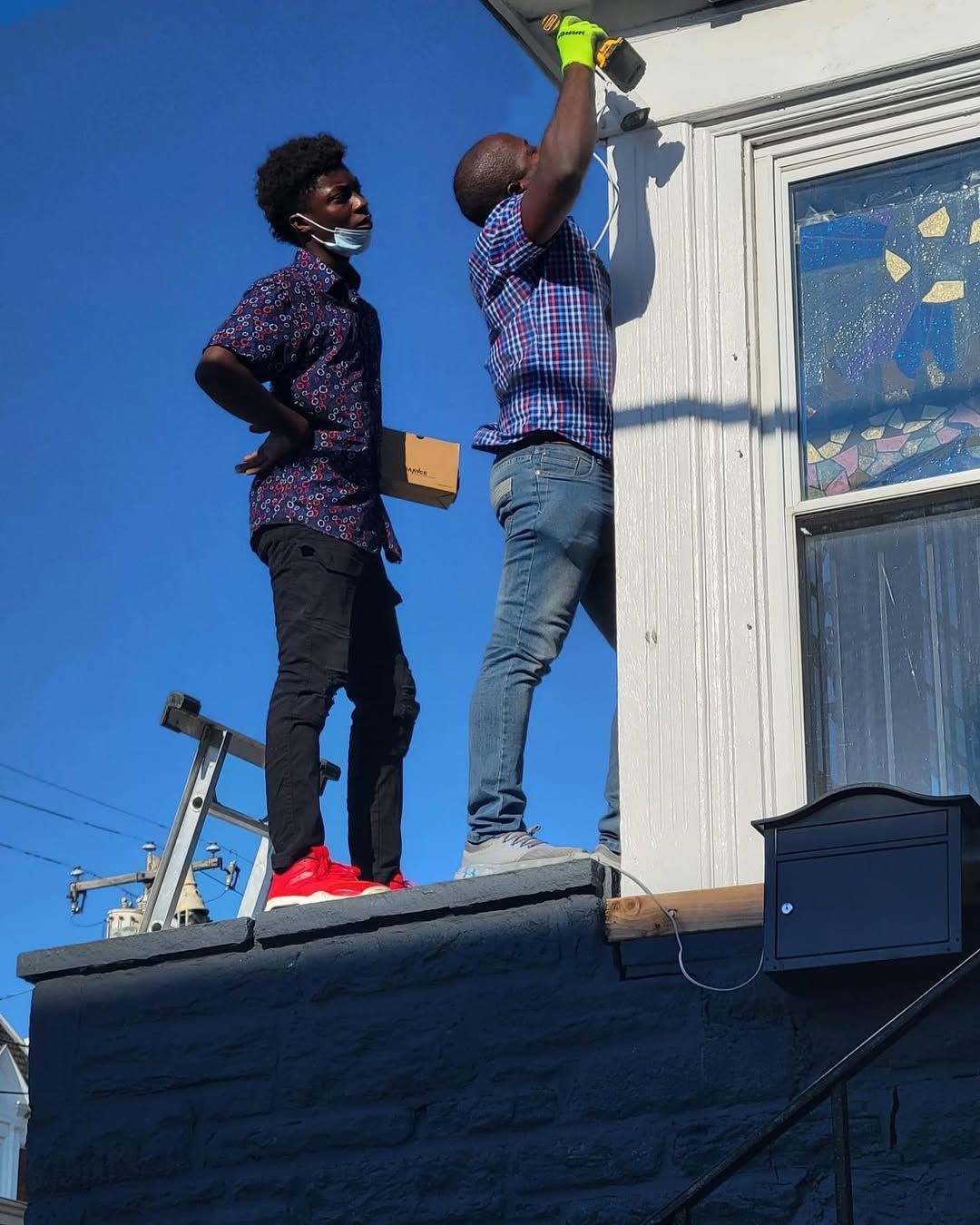 Two people painting an exterior wall on a sunny day. One paints, the other watches with a small box.