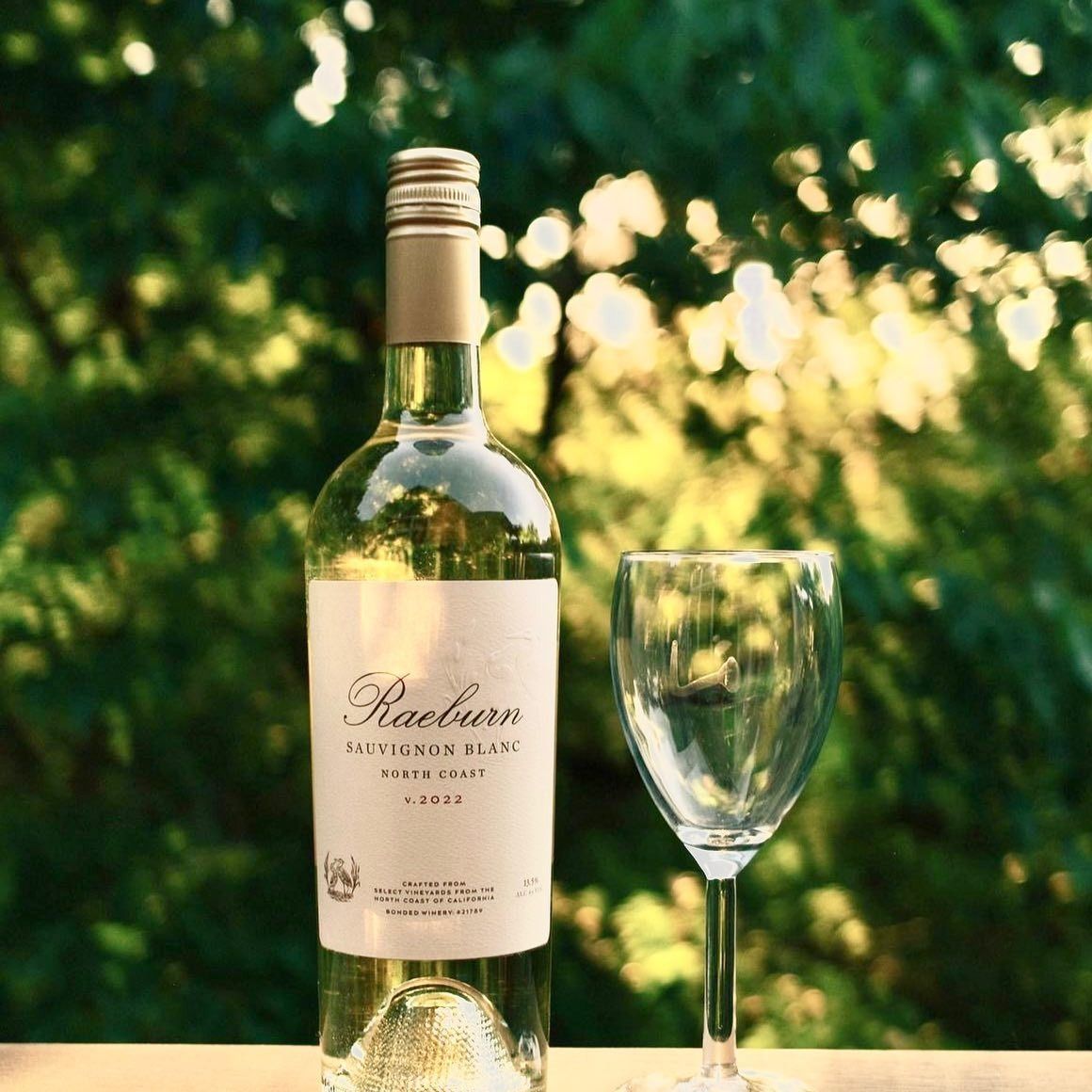 Bottle of white wine and empty glass on a table outside with trees in the background.