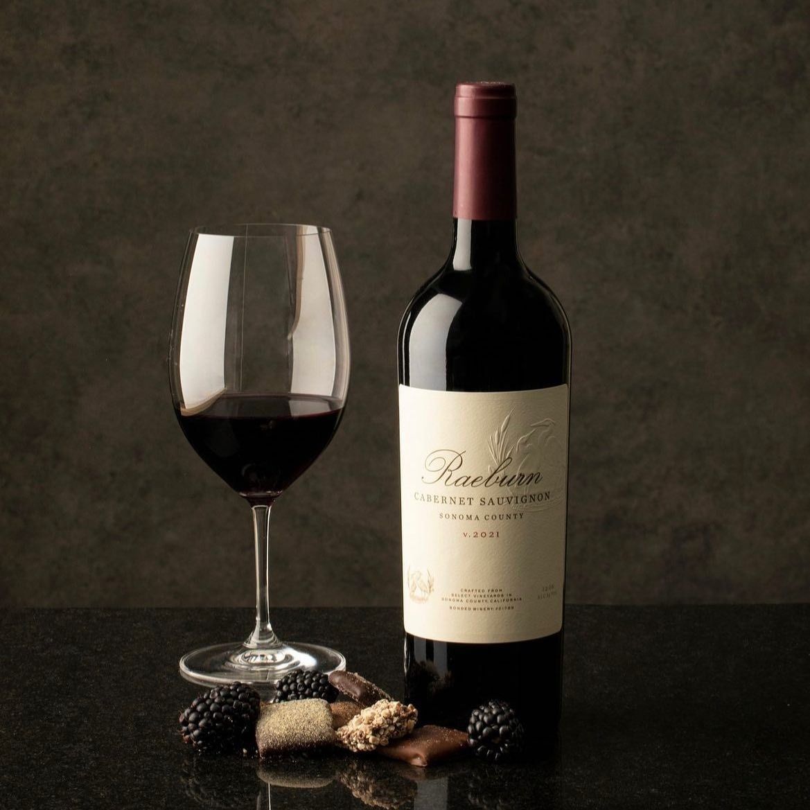 A bottle of red wine and a glass with red wine, along with a few chocolates and blackberries, set on a dark surface.