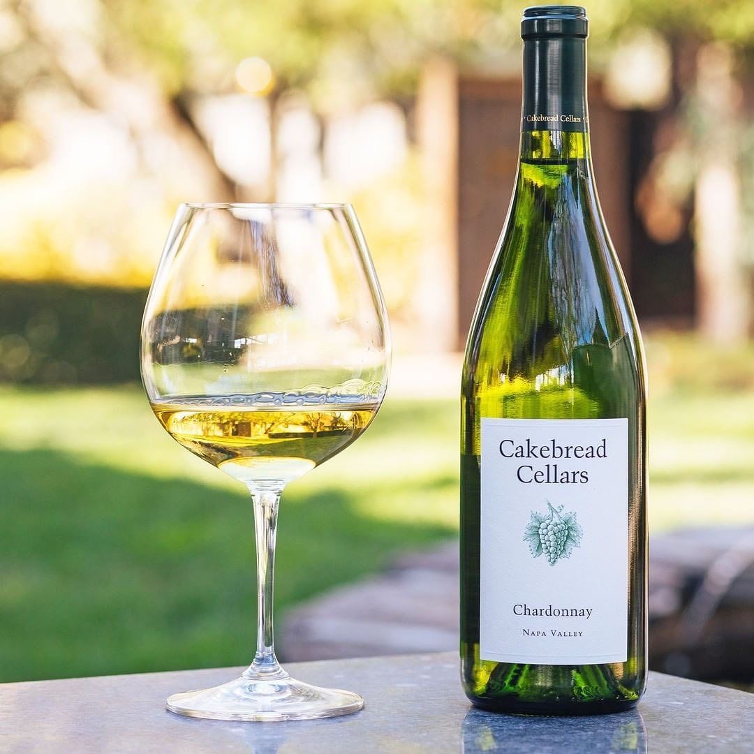 Bottle of Cakebread Cellars Chardonnay next to a wine glass filled with wine, on an outdoor table.