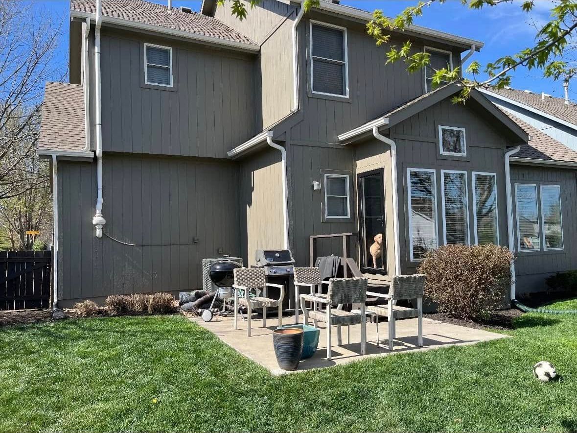 Backyard with gray house, patio, grill, chairs, dog in doorway, and green grass.