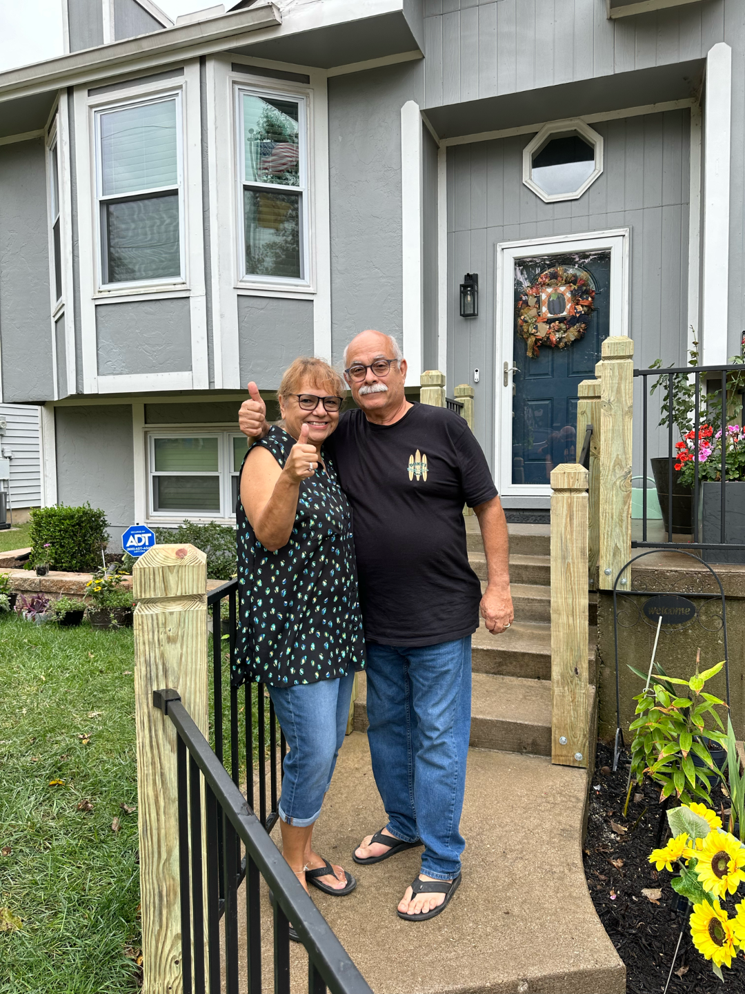 Couple smiling with thumbs up in front of a gray house with a dark blue door.