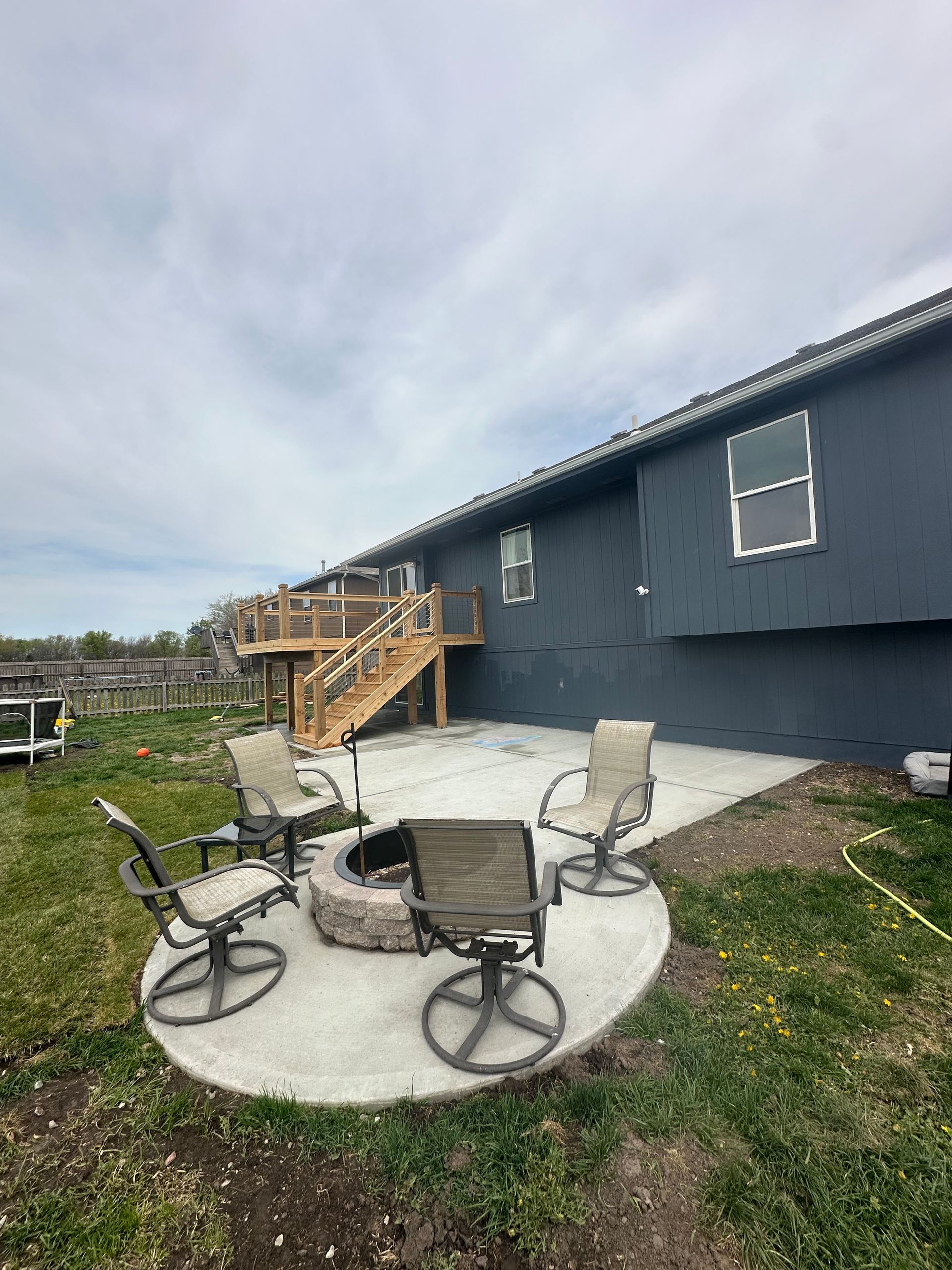 Concrete patio with fire pit, chairs, and wooden deck on a dark-painted house. Cloudy sky.
