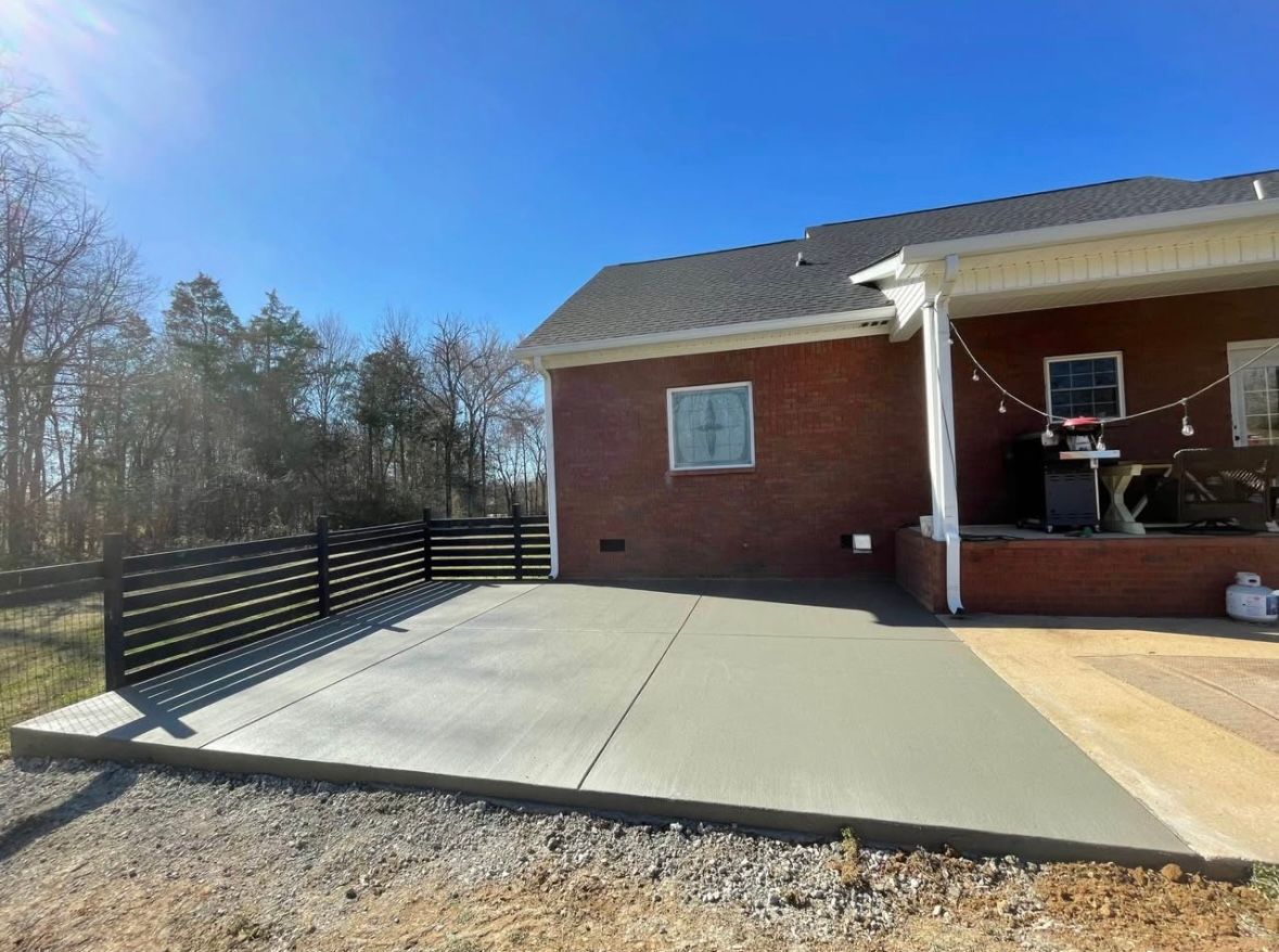Newly poured concrete patio next to a brick house, with a black fence and a sunny sky.