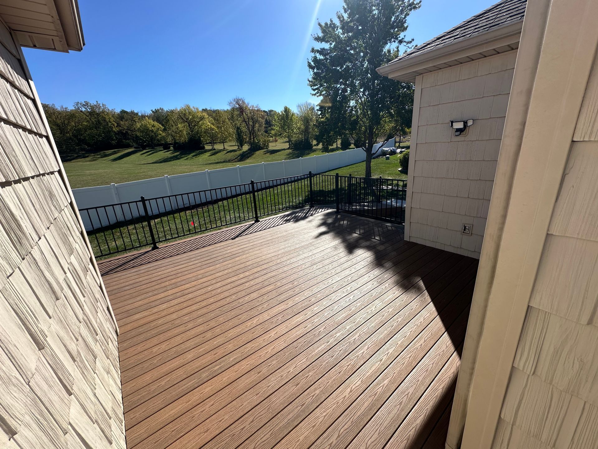 Wooden deck with a black fence overlooking a green field and trees, seen on a sunny day.