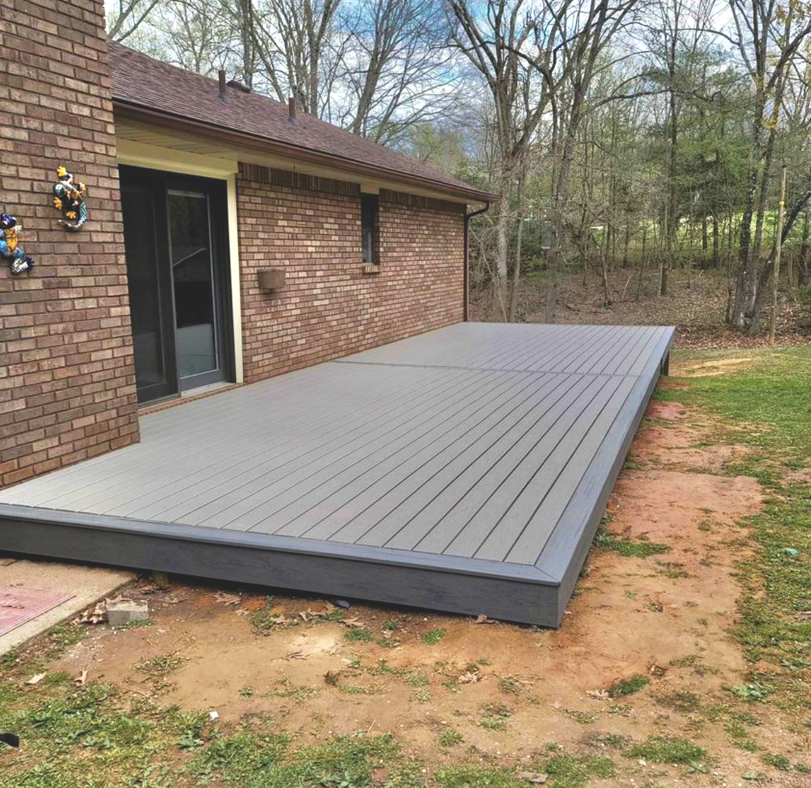 Gray composite deck attached to a brick house, surrounded by grass and trees.