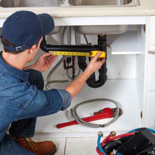 A man measuring a pipe under a sink with a tape measure