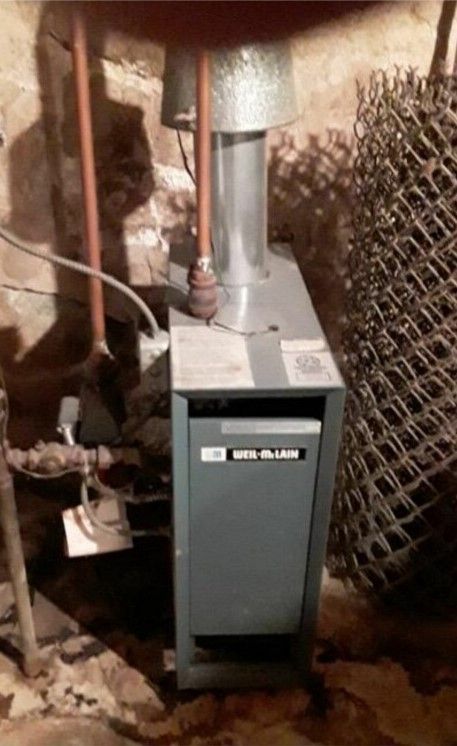 A gas boiler is sitting in a basement next to a wire fence.