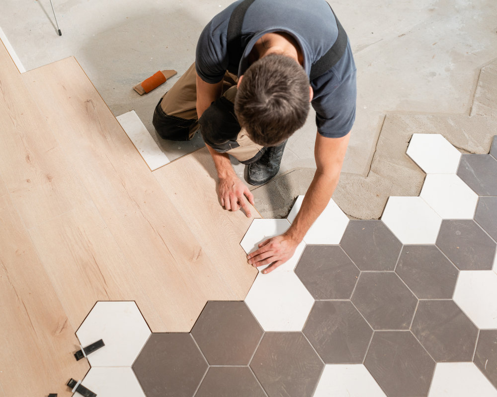 A man is laying tiles on a wooden floor.