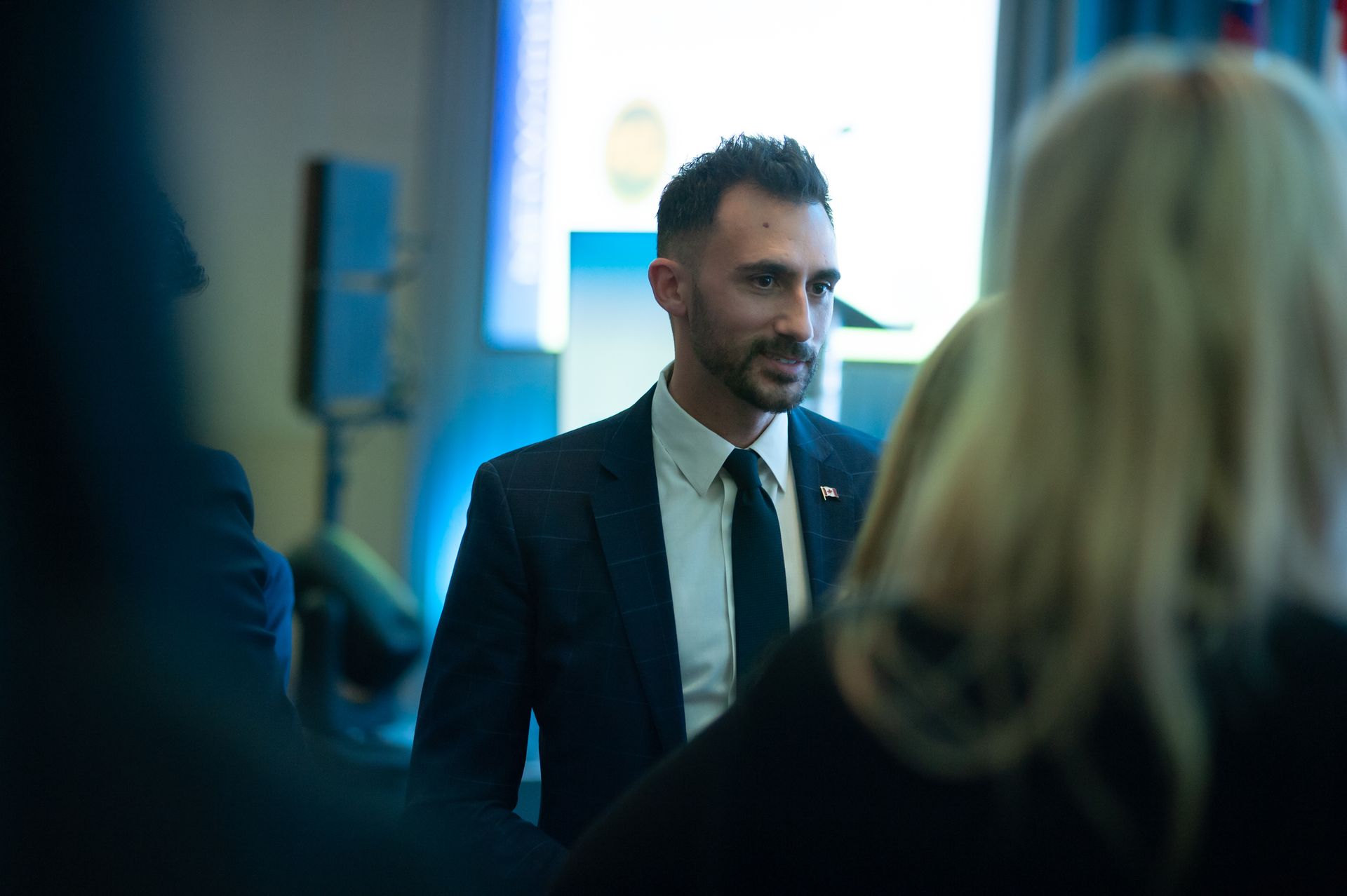 Minister Stephen Lecce attending an event. Interior setting with blurred background and people.