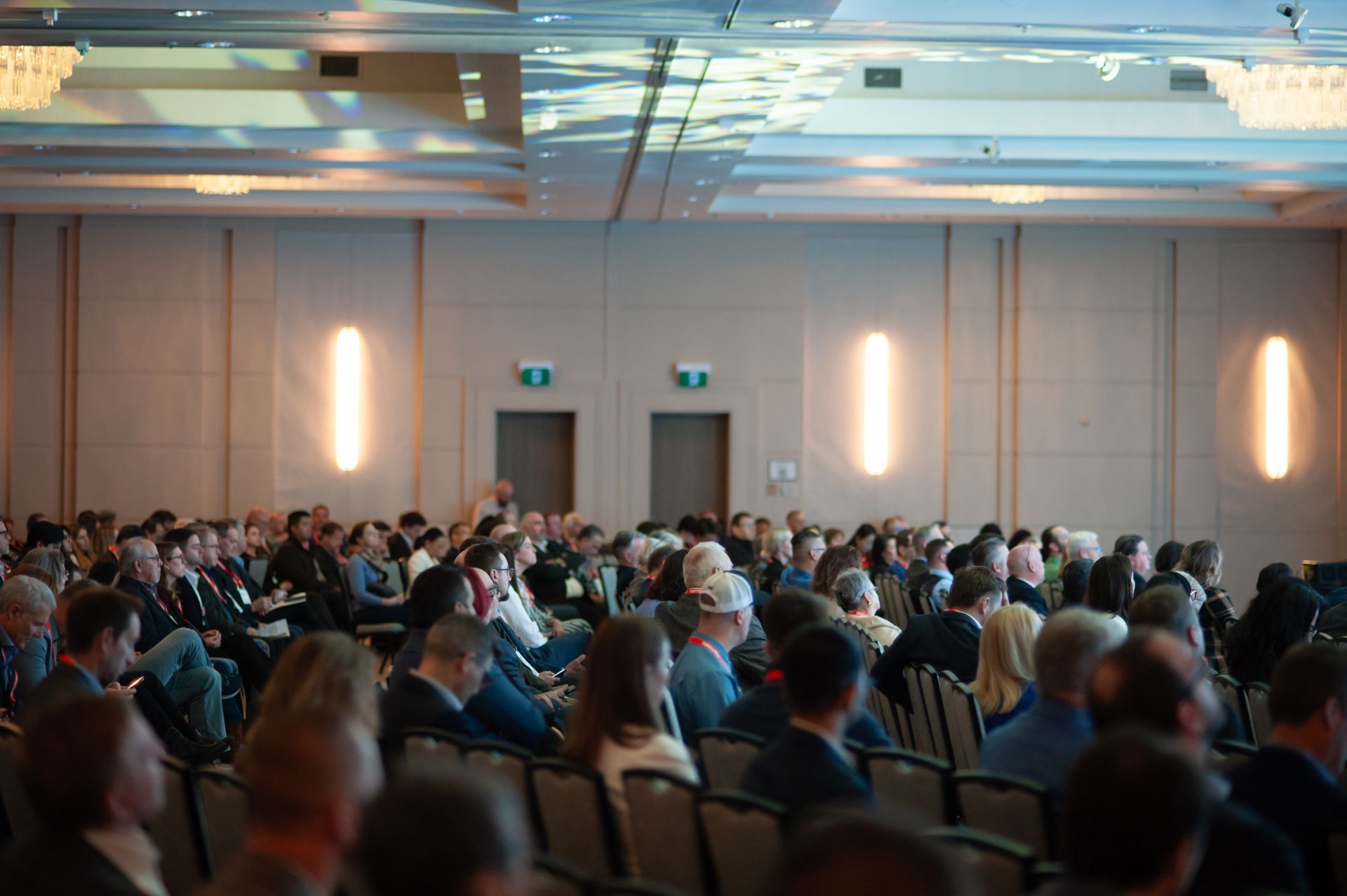 Audience in a conference room; beige walls, bright lights, people seated and listening.