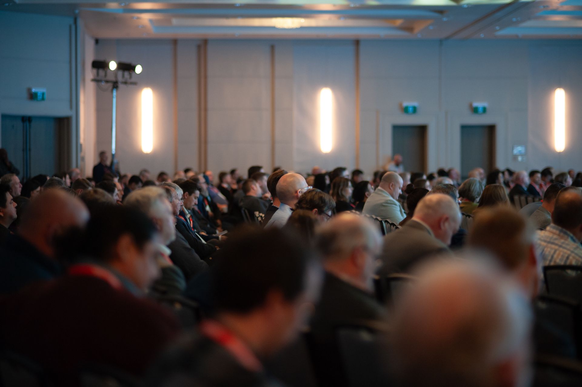 Audience seated in a large conference room, lit with dim blue and warm white lights.
