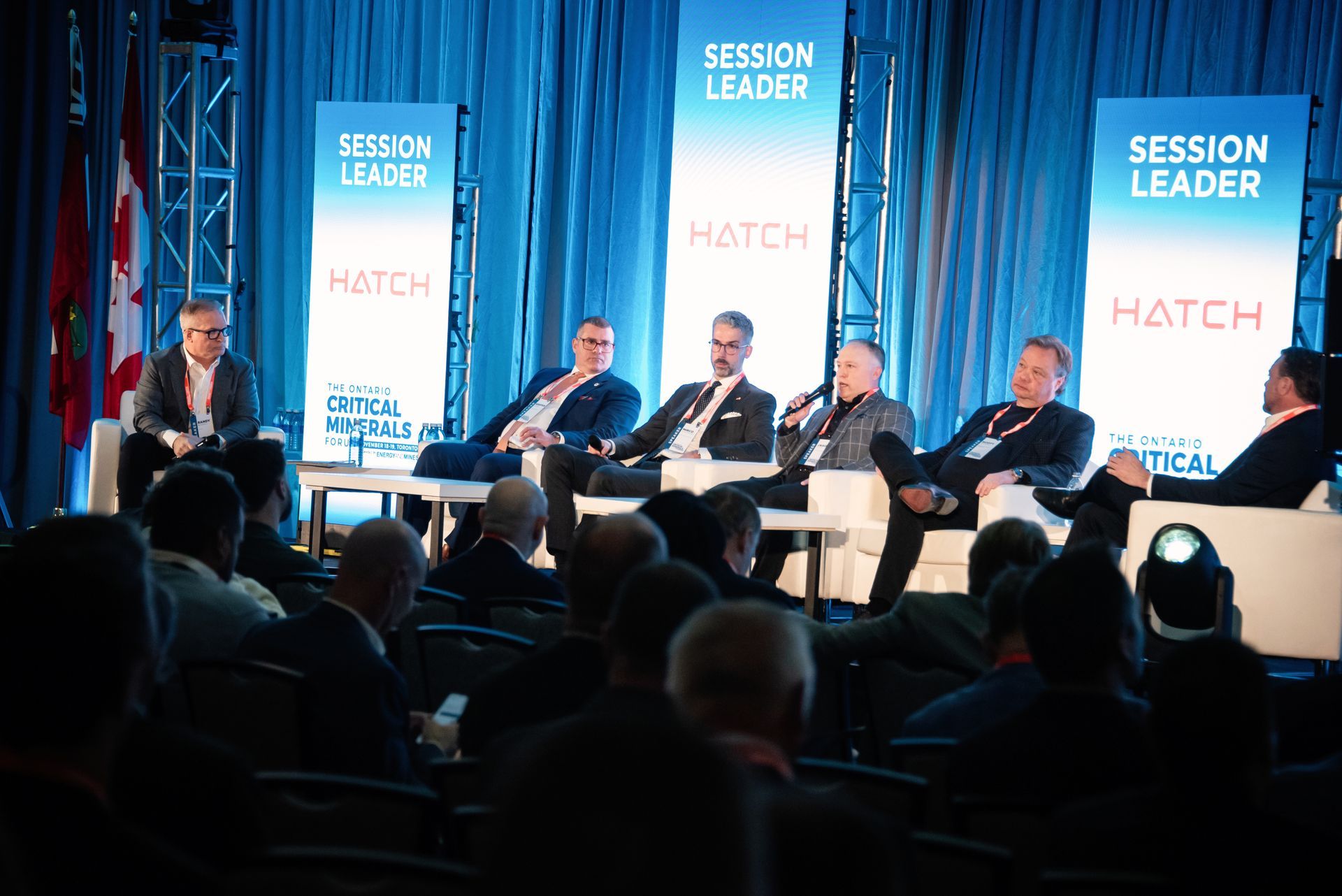 Panel of men sitting on stage, speaking at a conference, with an audience in front.