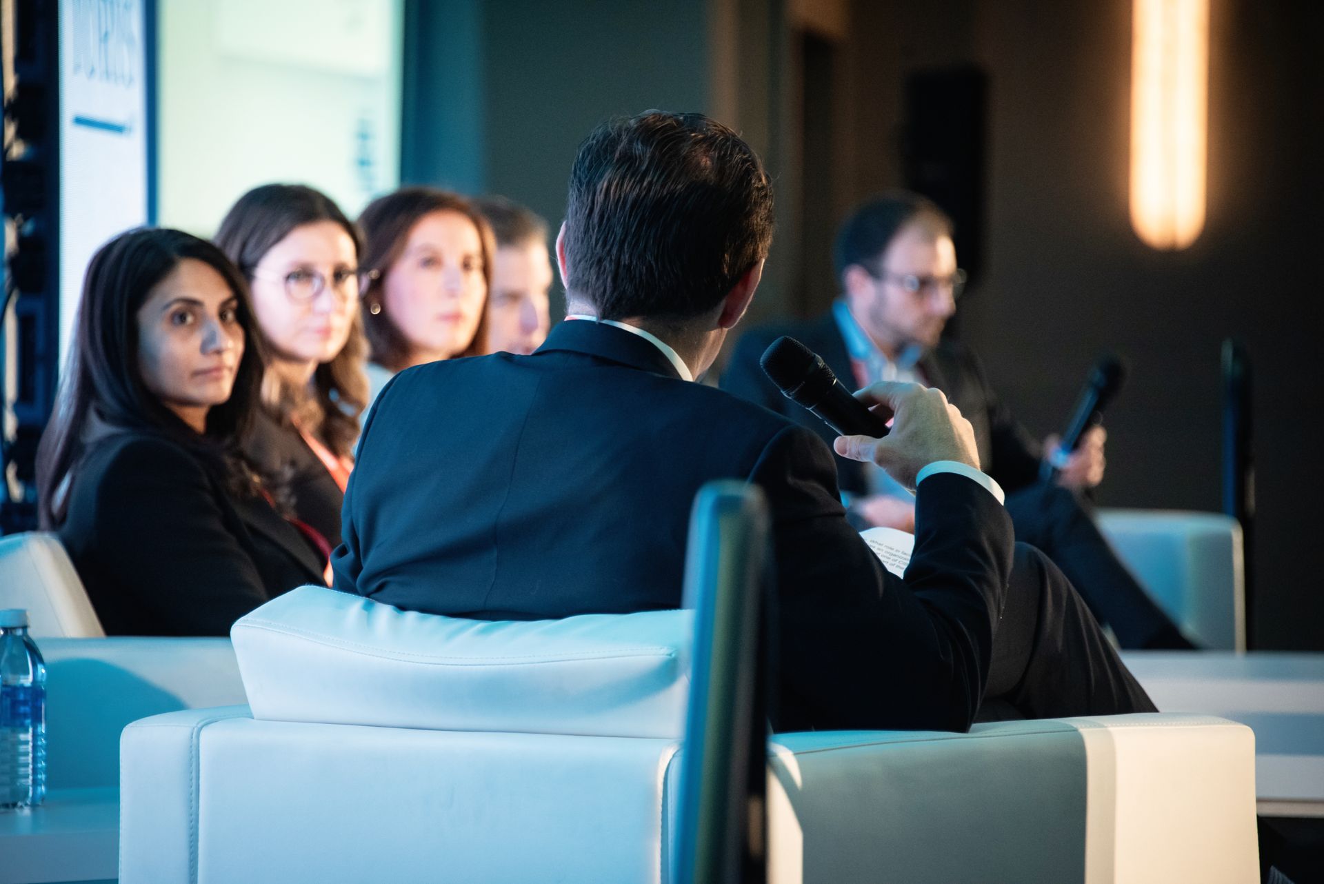 Man speaking into a microphone, facing a panel seated on white sofas. People listen.