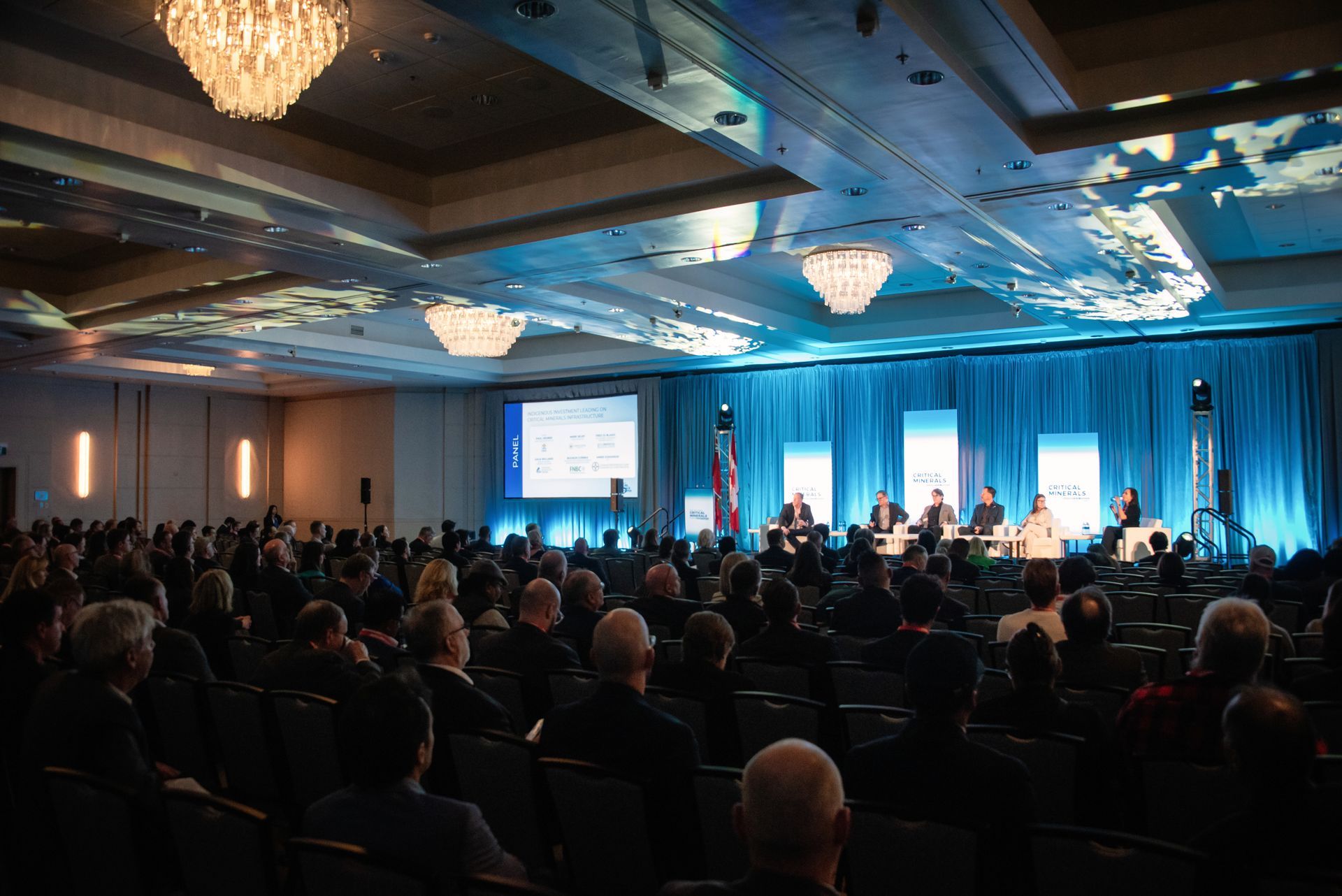 Conference in a large room, panelists on stage, audience seated facing forward. Blue and white lighting.