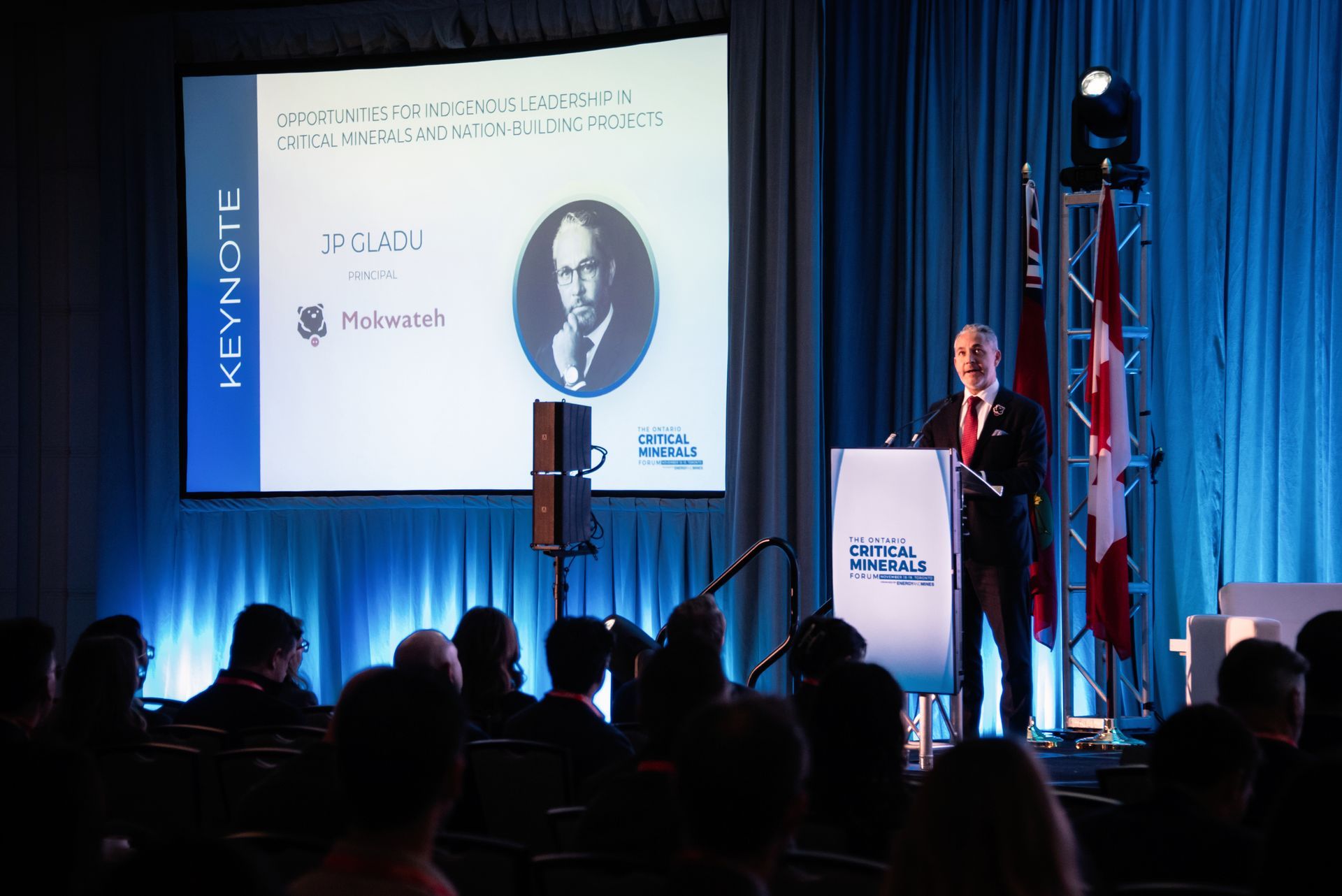 Man speaking at a conference keynote. Screen displays a portrait and title. Audience is seated.
