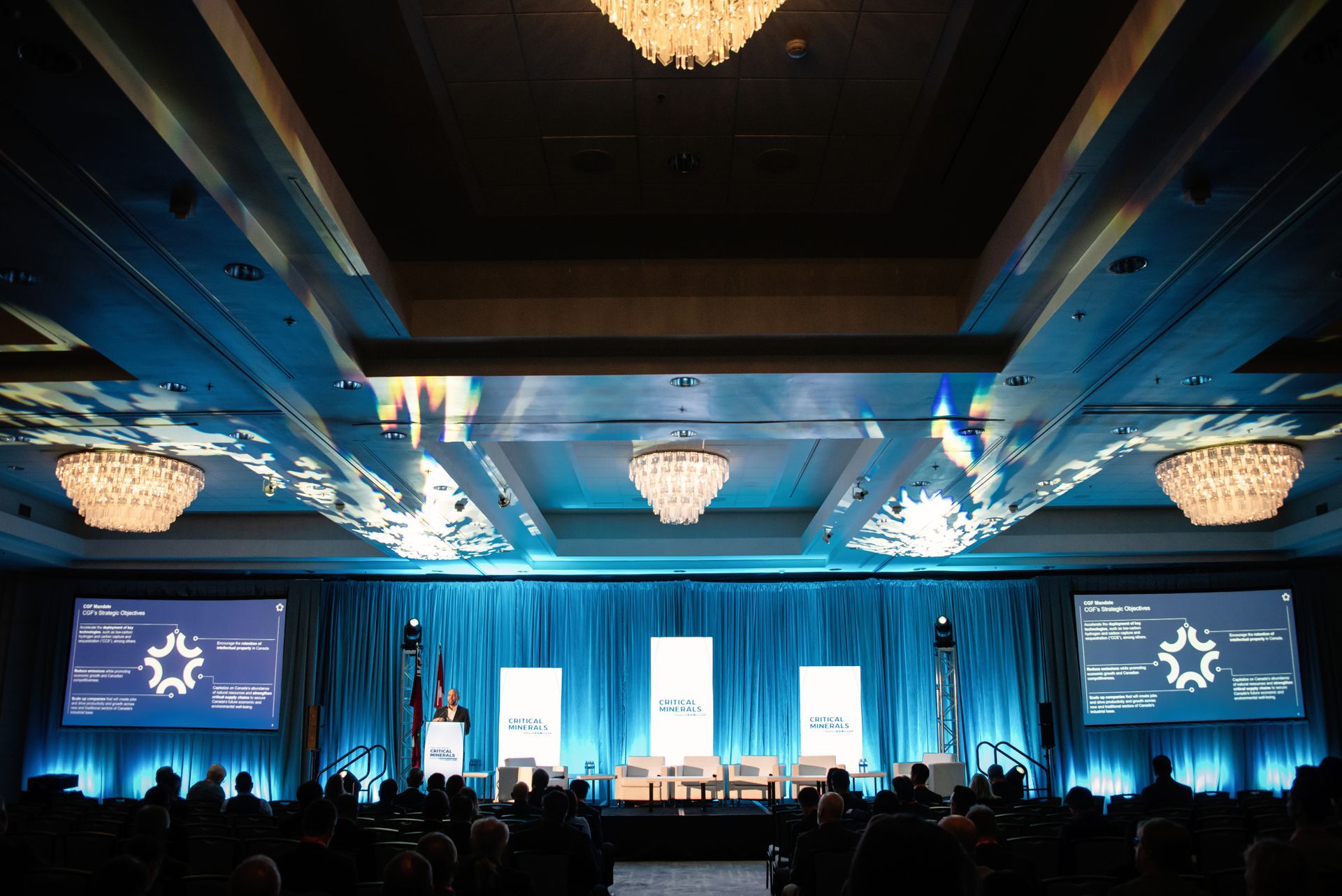Conference stage with presenters, audience, and blue lighting.