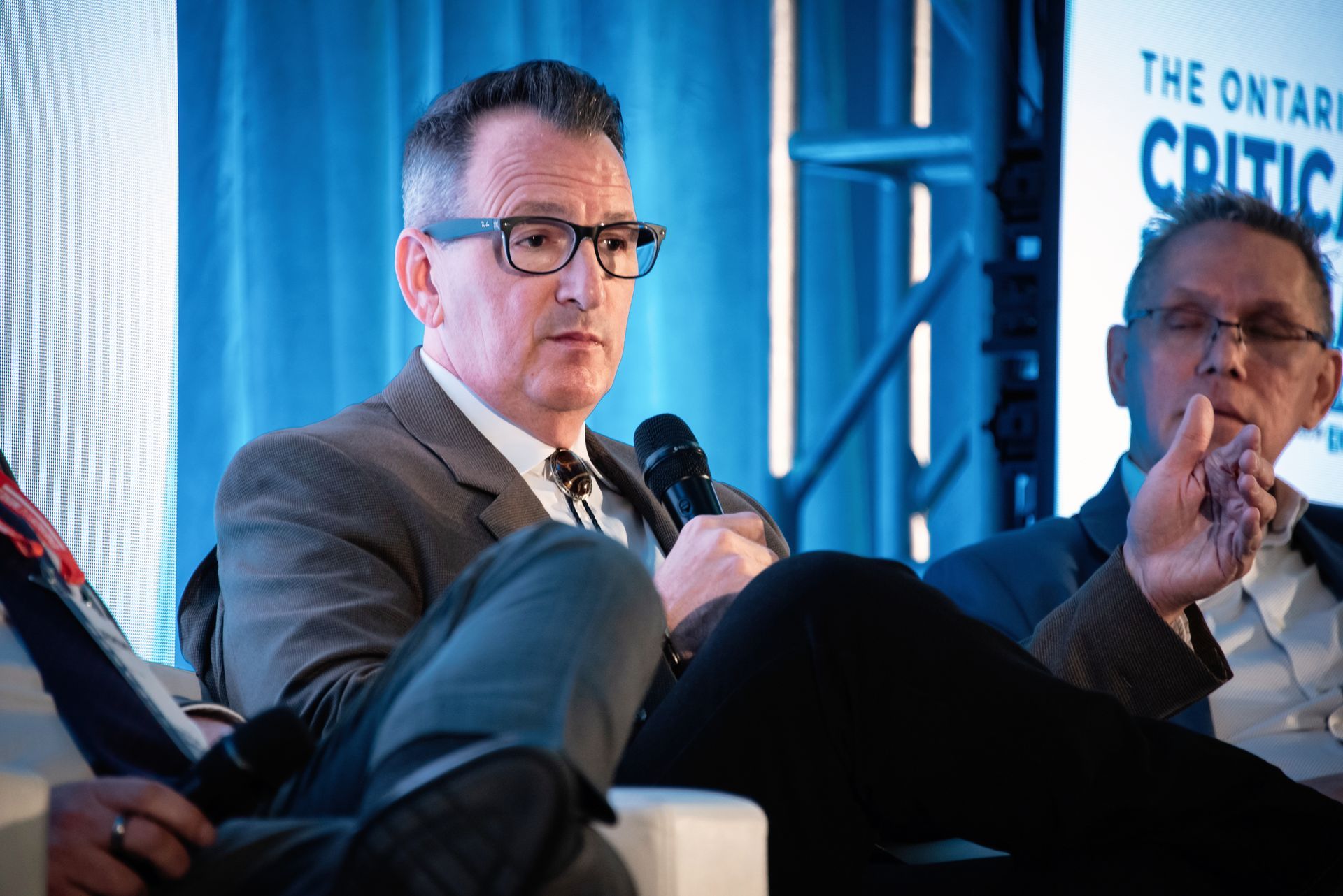 Minister Rickford speaks into microphone, seated at a conference table with another man.