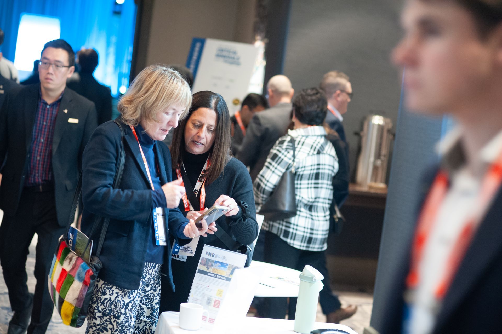 Two women looking at a phone at a conference table, other attendees in background.