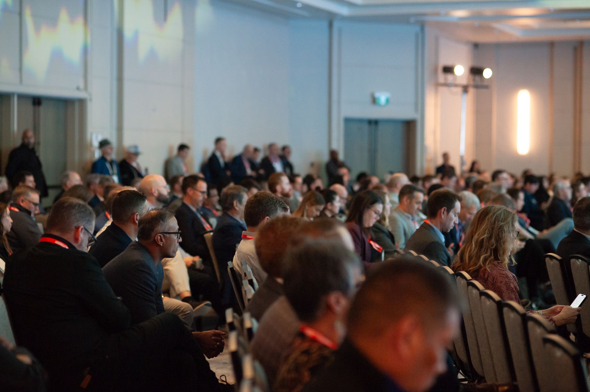 Audience seated in a conference hall, listening. Some visible from shoulders up. Brightly lit room.