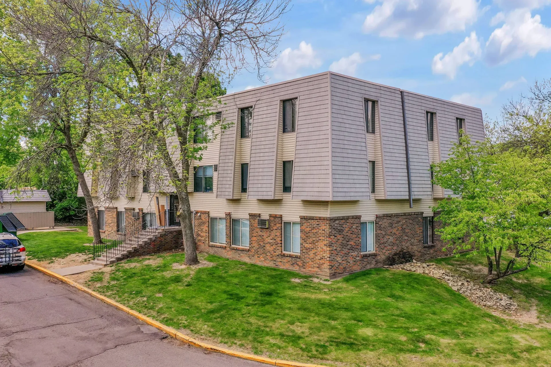 Exterior view of a multi-family apartment building with brick lower level, beige siding, and surrounding trees.