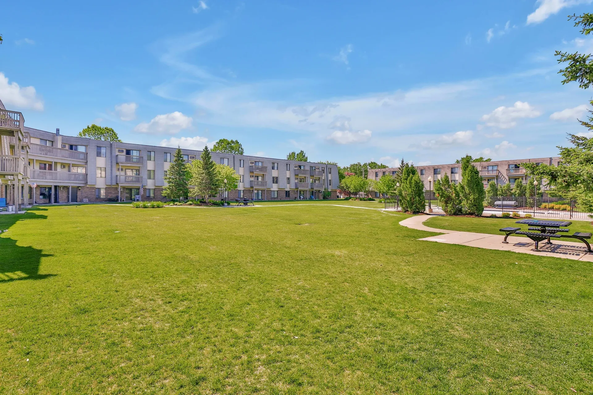 Wide view of a grassy courtyard surrounded by apartment buildings, with benches and trees under a blue sky.