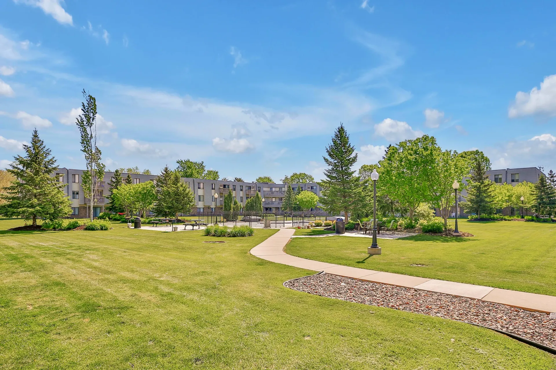 Wide view of a well-kept apartment community lawn with a curved walkway, benches, and buildings in the background.