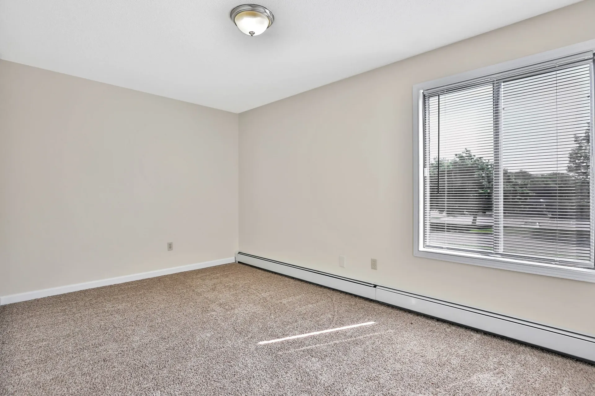 Empty beige apartment room with window and blinds, carpeted floor.