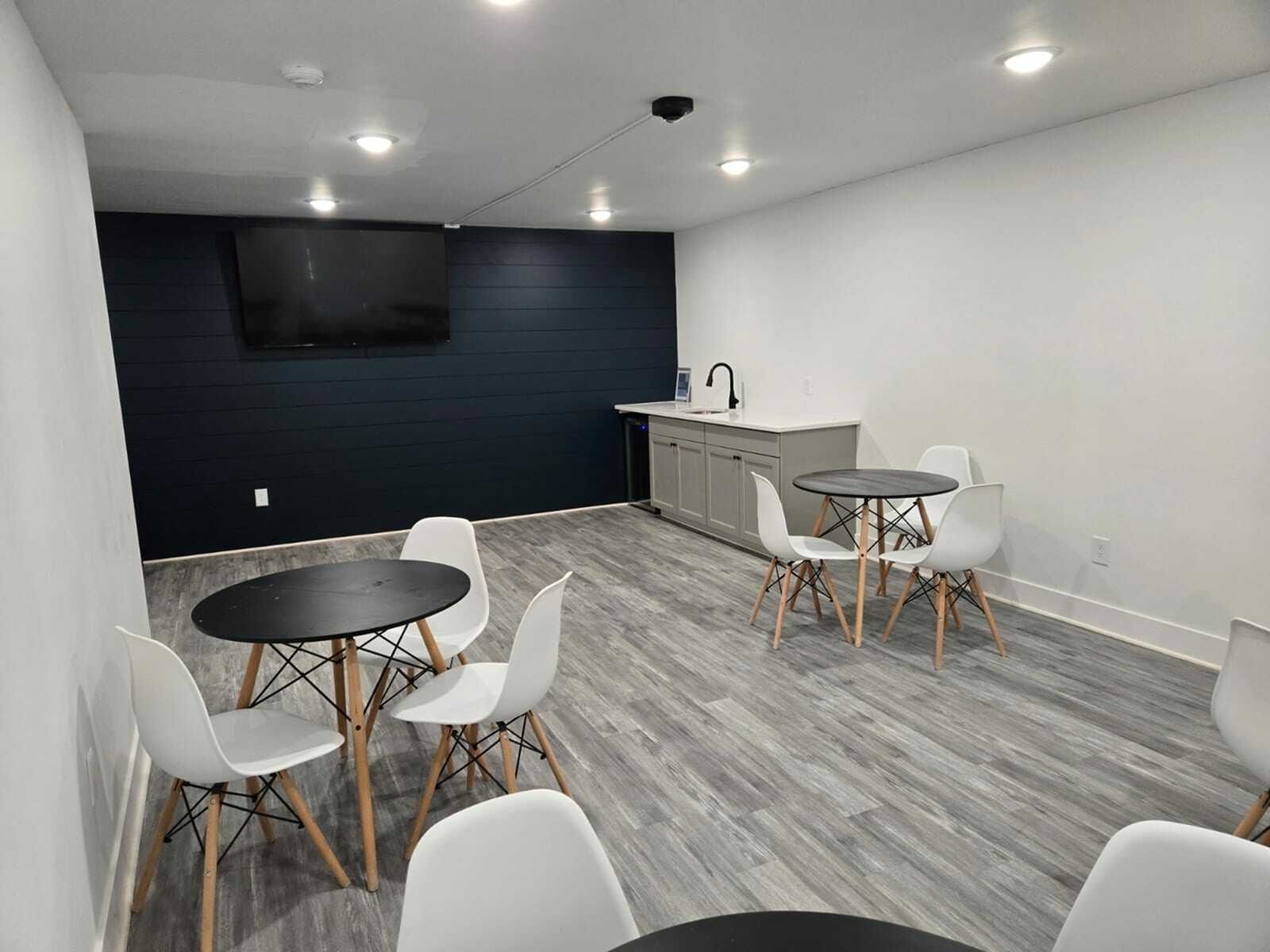 Indoor communal lounge with round black tables, white chairs, a kitchenette, and a wall-mounted TV.