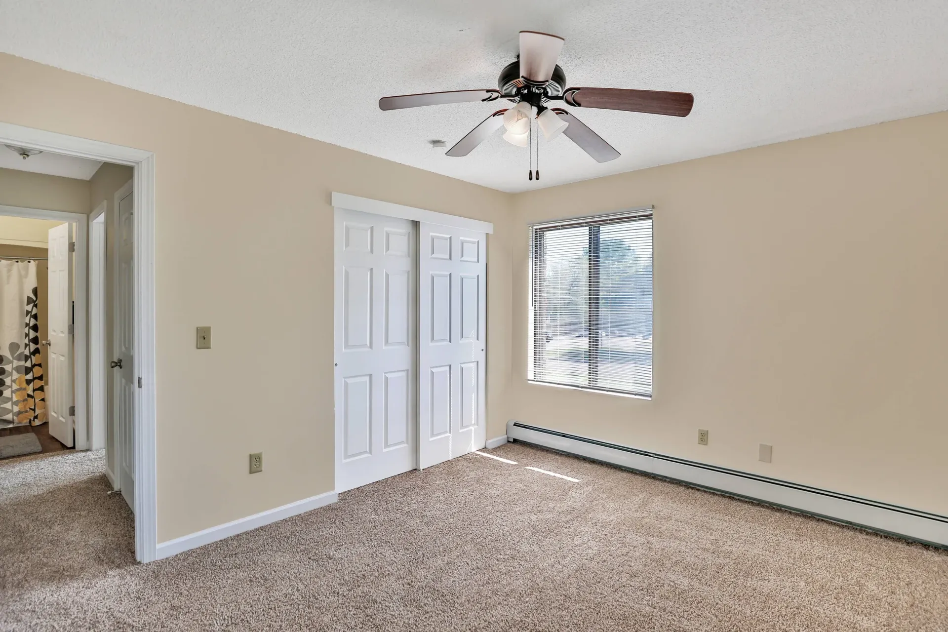 Interior bedroom with beige walls, ceiling fan, double closet doors, and a window with blinds.