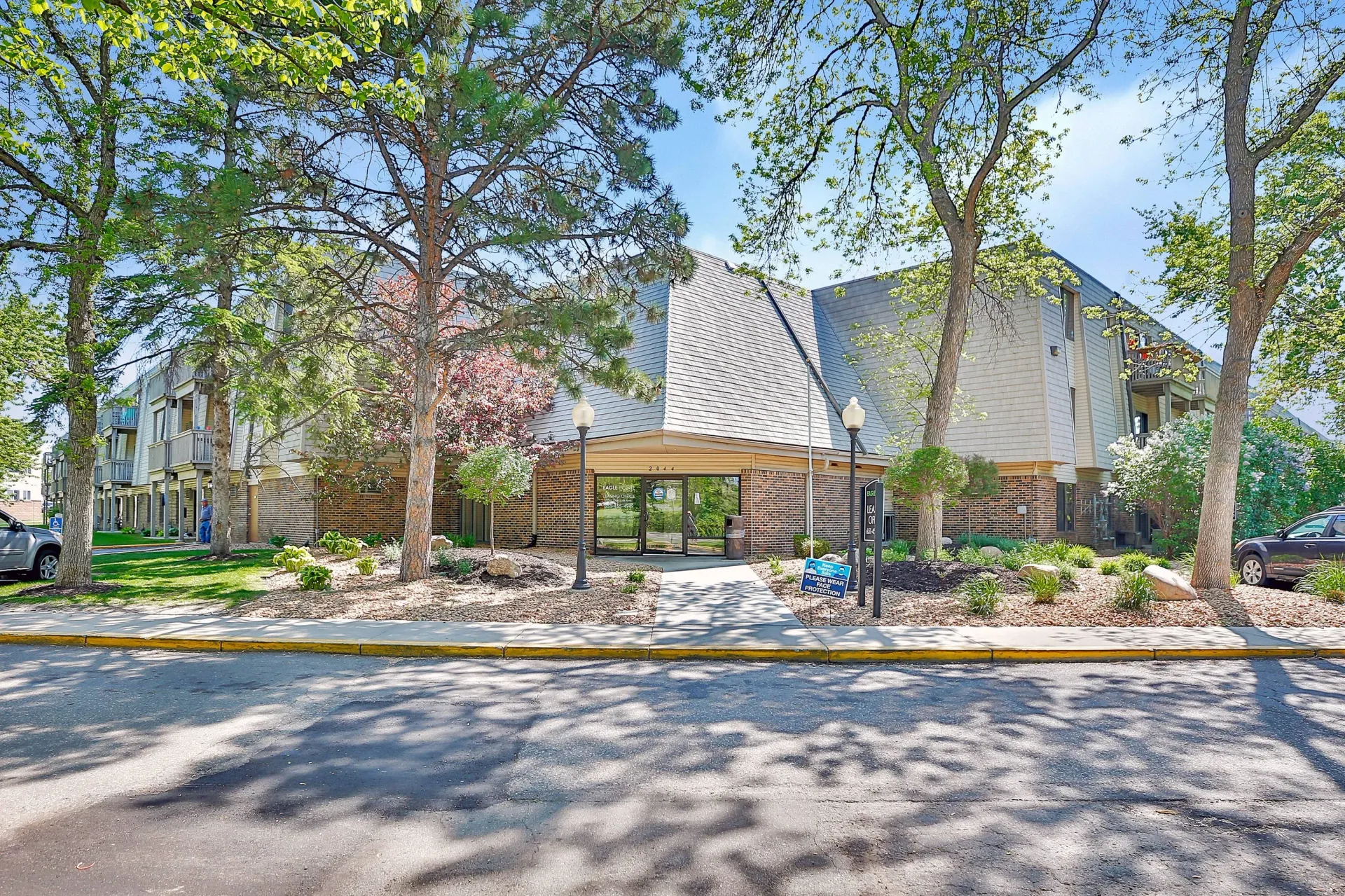 Exterior view of a multifamily community entrance with brick base, glass doors, and surrounding trees.