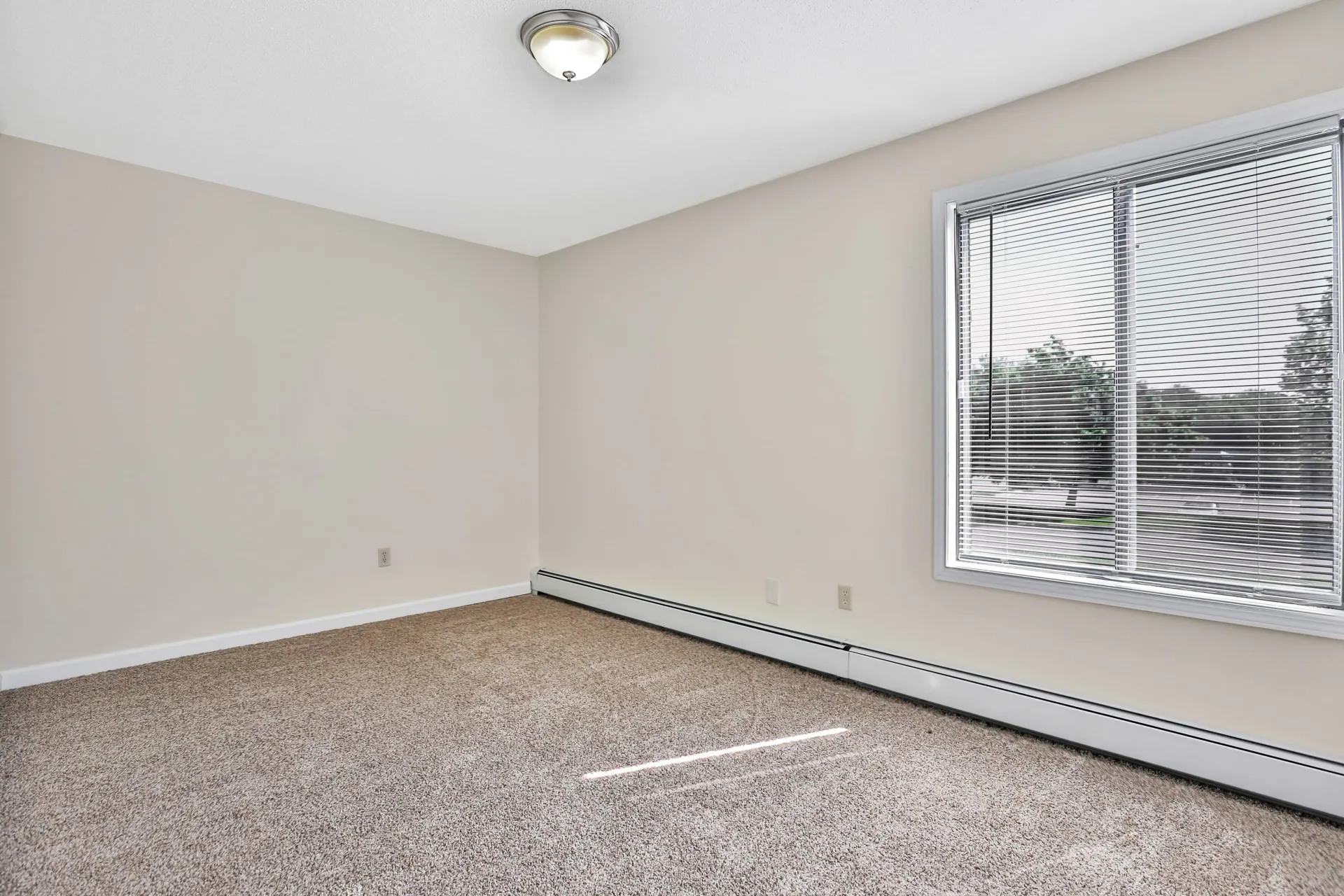 Empty room with tan walls, carpet, and a window with blinds at Eagle Point Apartments in West St. Paul, MN.