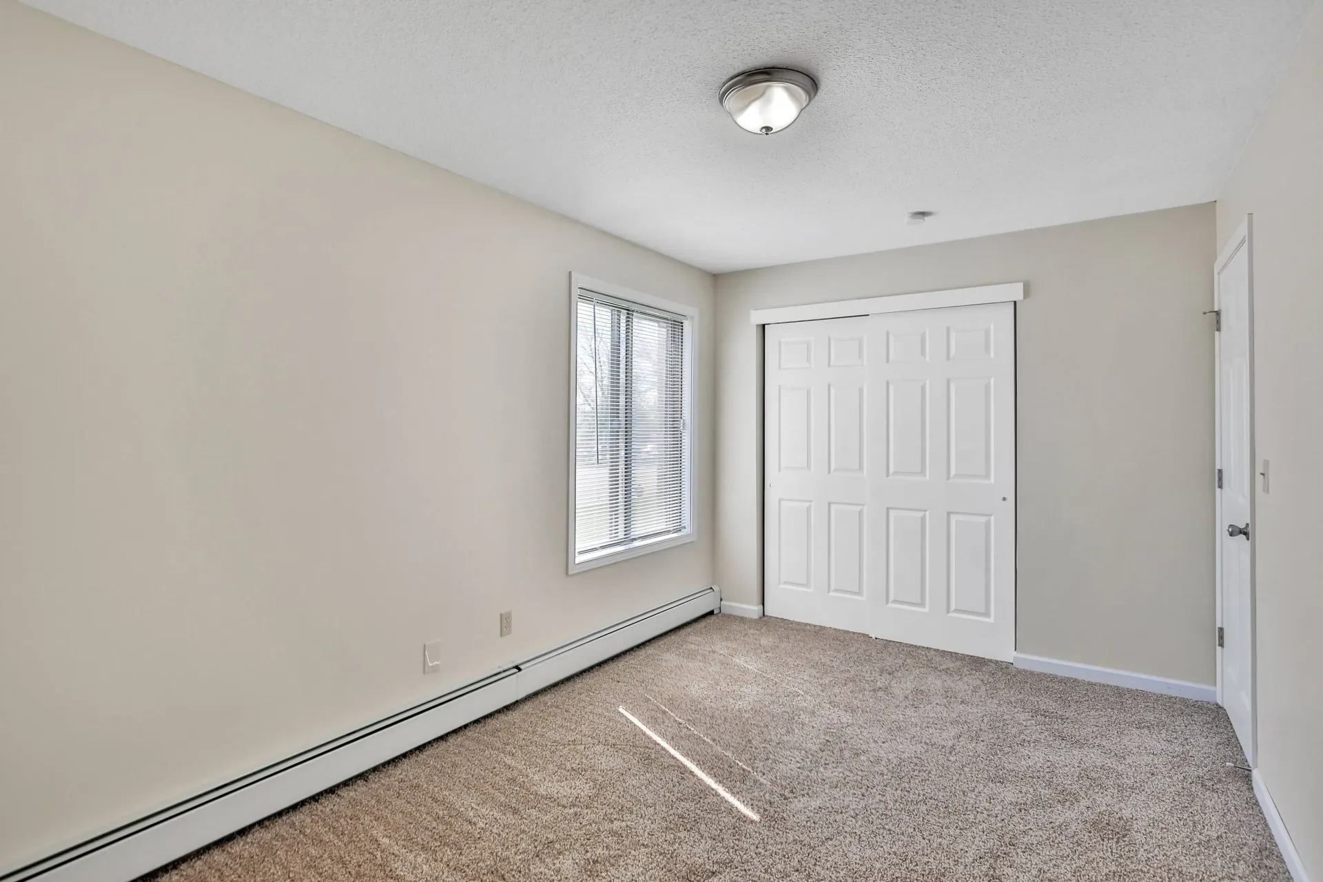 Empty bedroom with beige walls, carpet, sliding closet doors, and a window at Eagle Point Apartments in West St. Paul, MN.
