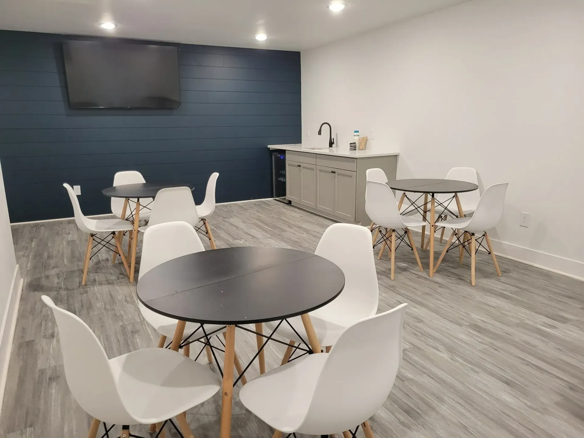 A modern lounge with round tables, white chairs, a wet bar, and a mounted TV on a blue wall at Eagle Point Apartments in West St. Paul, MN.