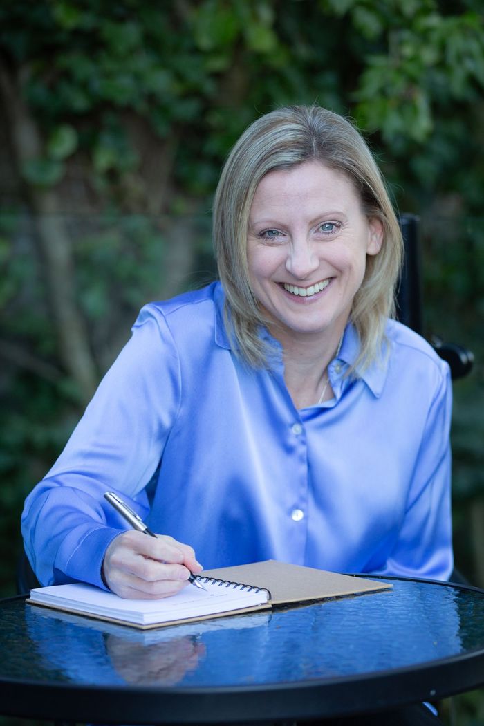 Picture of lady with blonde hair, smiling, holding a pen in hand and wearing a blue blouse