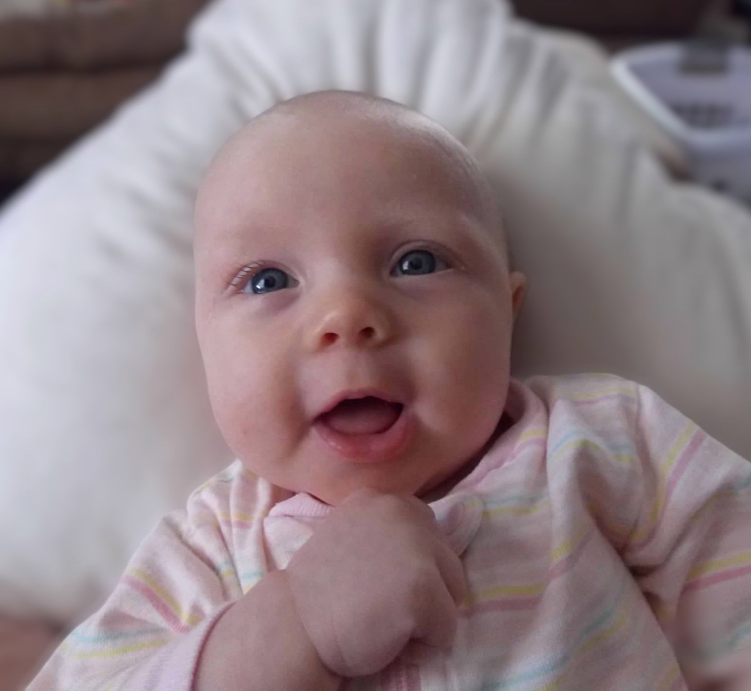 Smiling baby with blue eyes and bald head, wearing a pink striped onesie, lying against a white pillow.