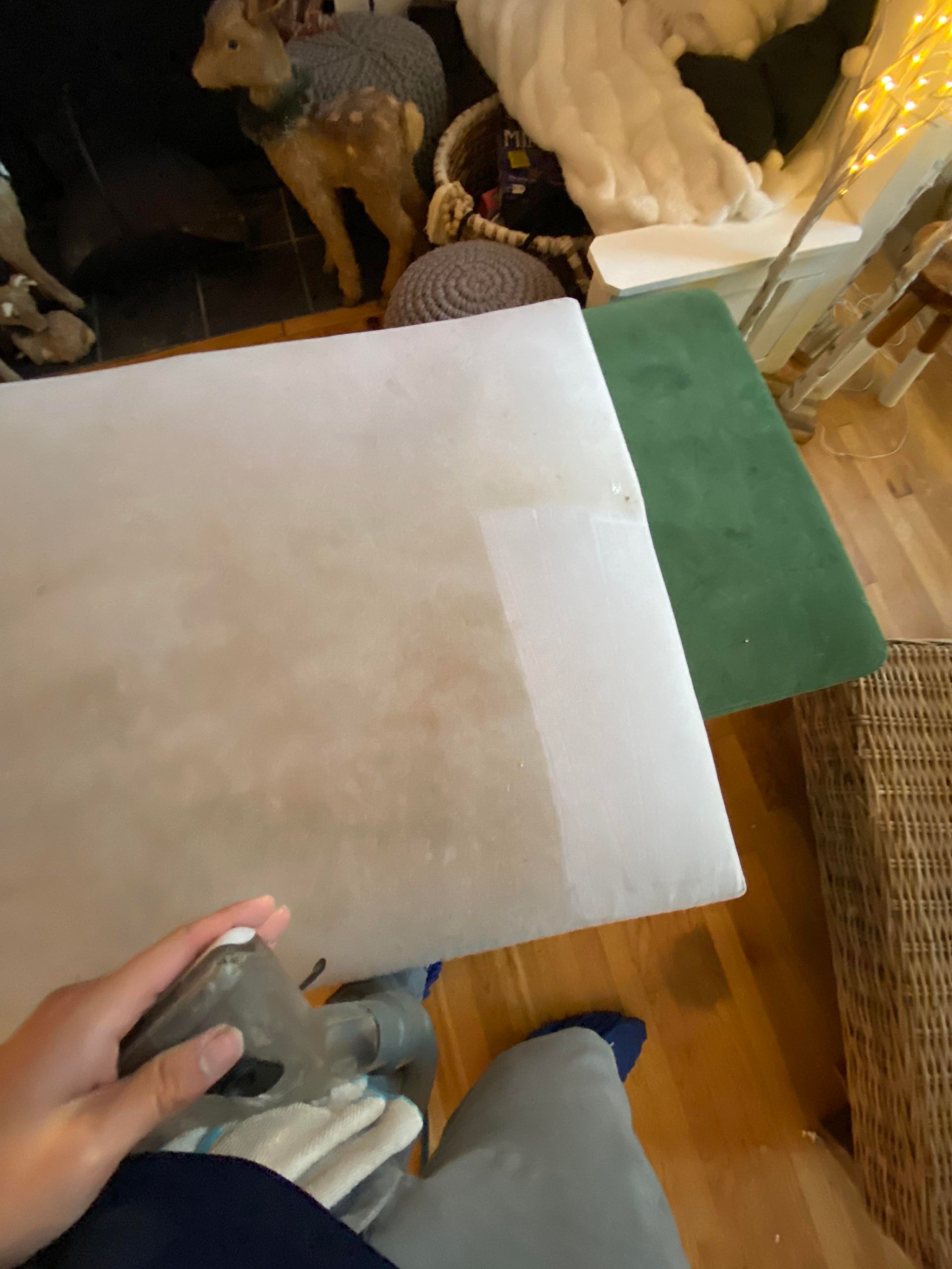 Person cleaning a light-colored cushion with a handheld cleaner. Adjacent is a green cushion. Wooden floor.