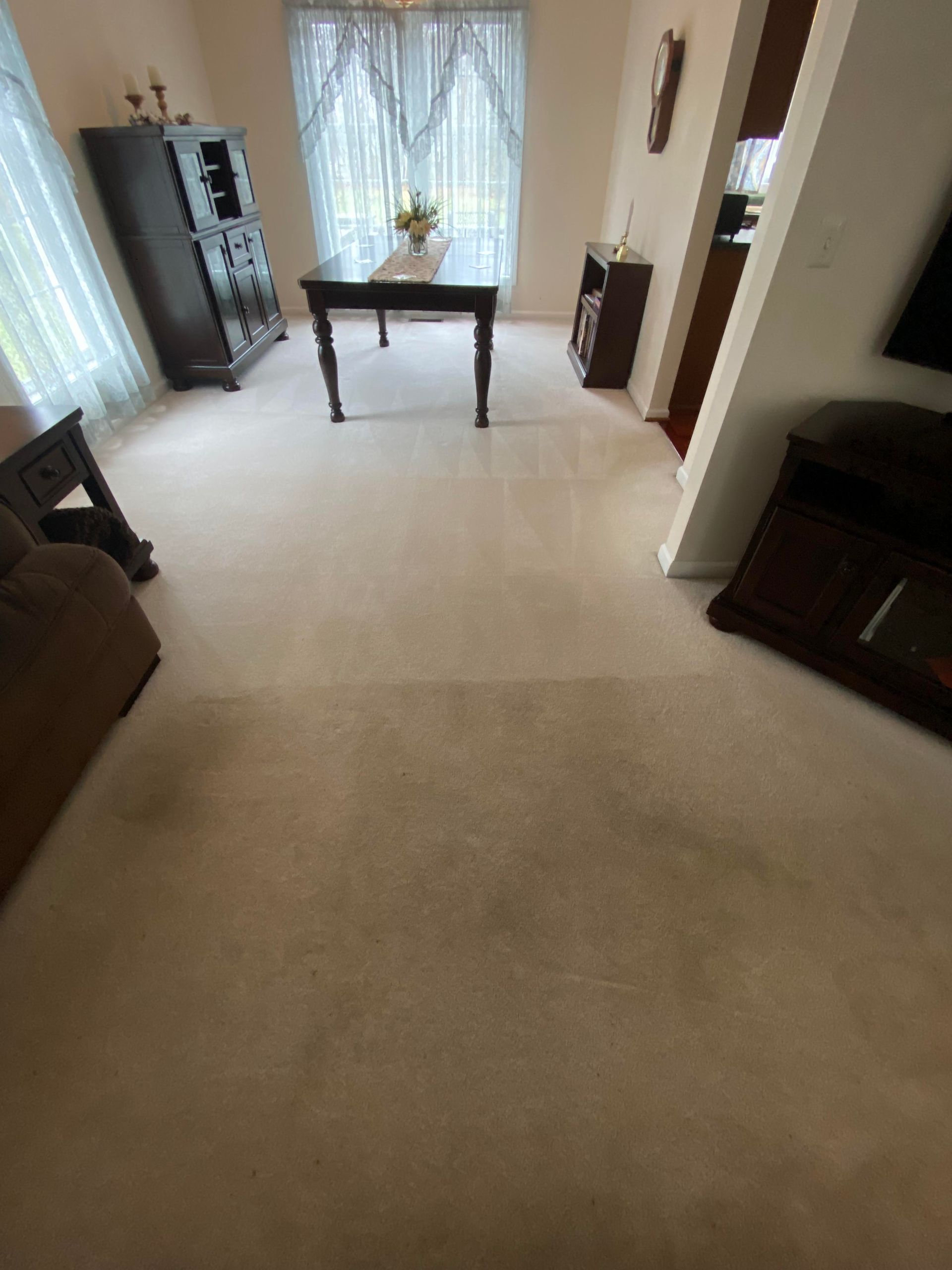 Beige carpeted room with dining table, dark wood furniture, and a window with sheer curtains.
