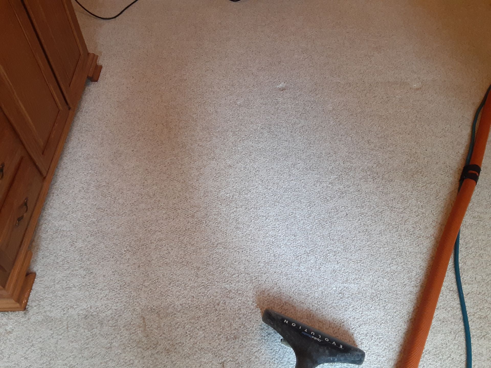 Tan carpet being vacuumed with a black and orange vacuum. A wooden cabinet is on the left.