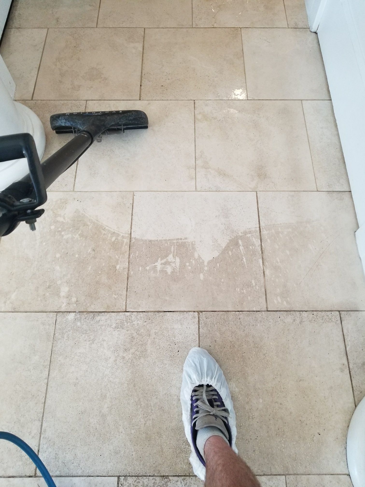 Person cleaning beige tile floor with a machine; one shoe is visible.