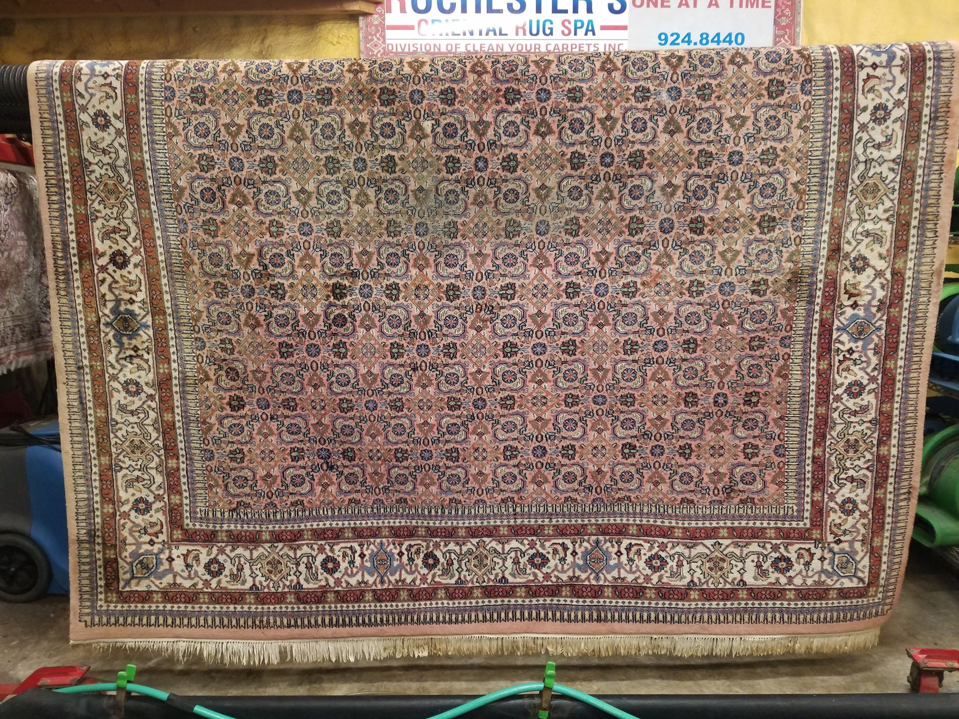 An ornate, rectangular rug with a patterned design, hanging on display. It features brown, beige, and red tones.