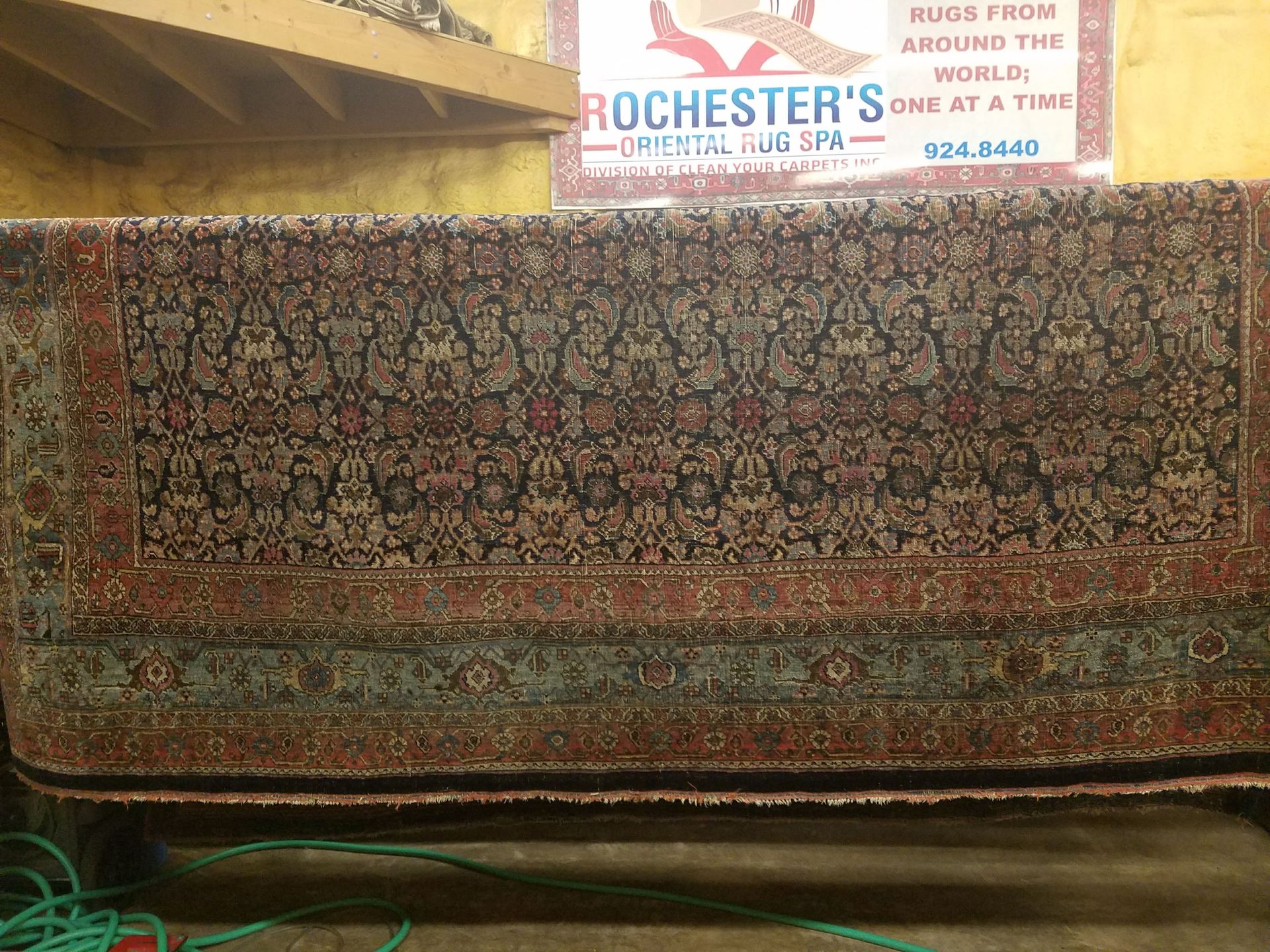 Rug at a rug dark central field with floral design, red and green borders, sign above reads 