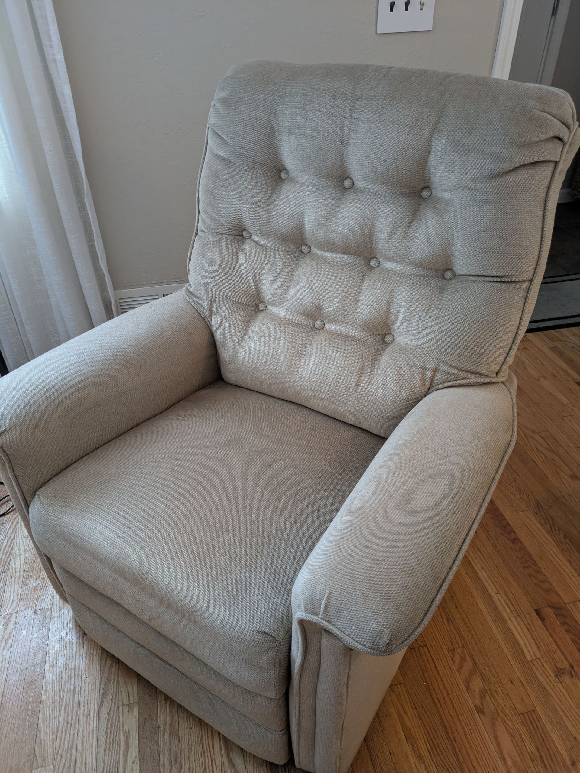 Beige upholstered recliner chair with button-tufted back, in a room with hardwood floors.