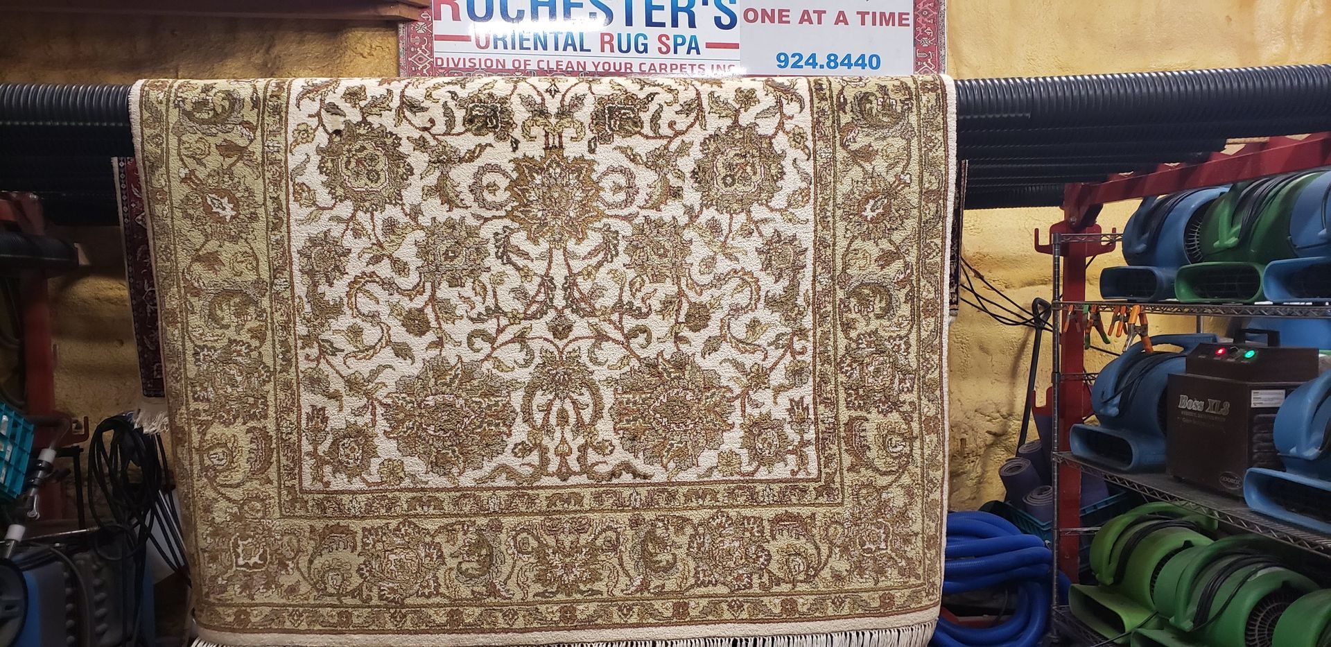 A light-colored rug with ornate floral designs hanging in a shop, with equipment in the background.