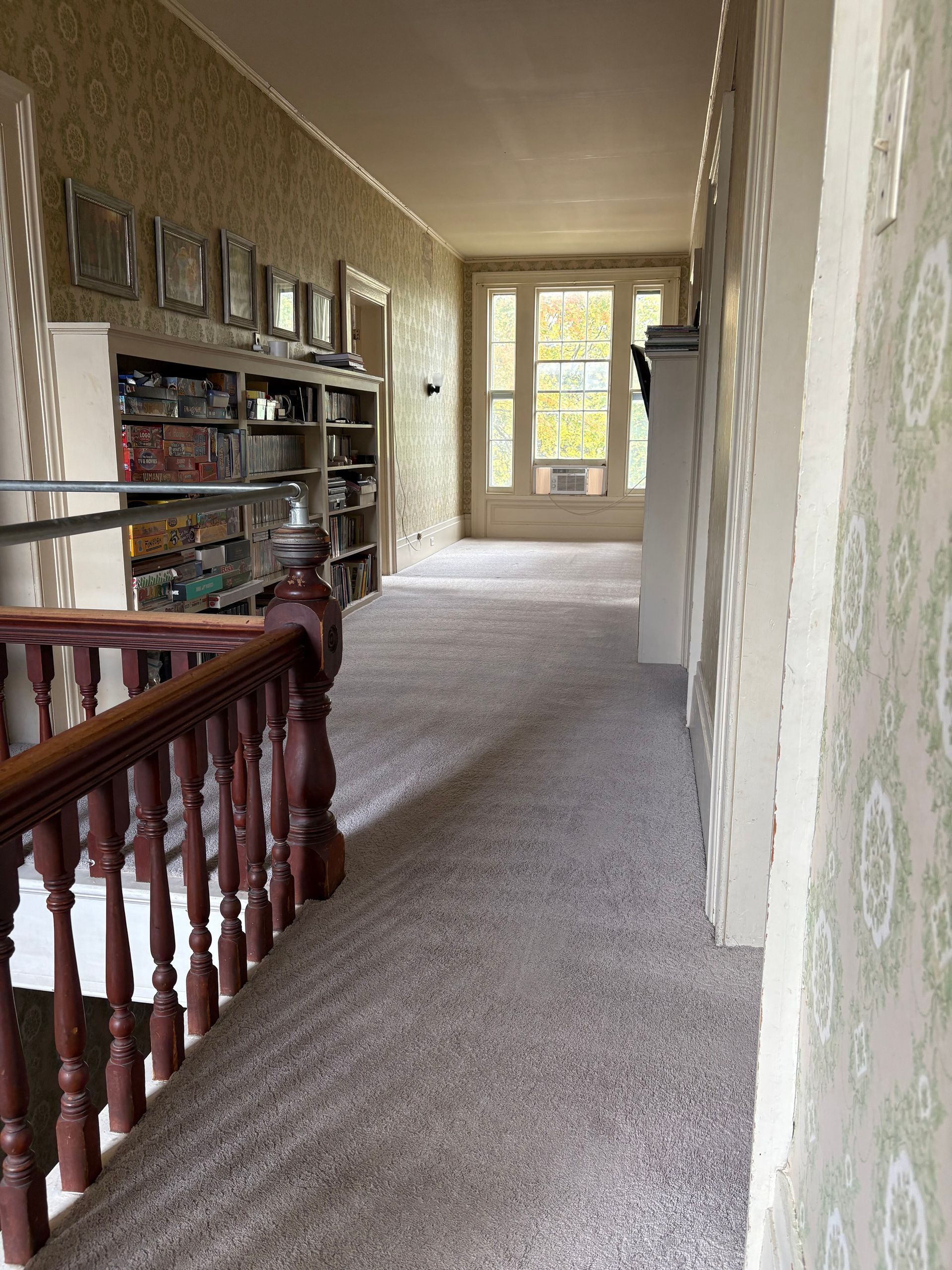 Hallway with bookshelves, staircase, and a window at the end. Carpeted floor, light-colored walls, and a wooden banister.