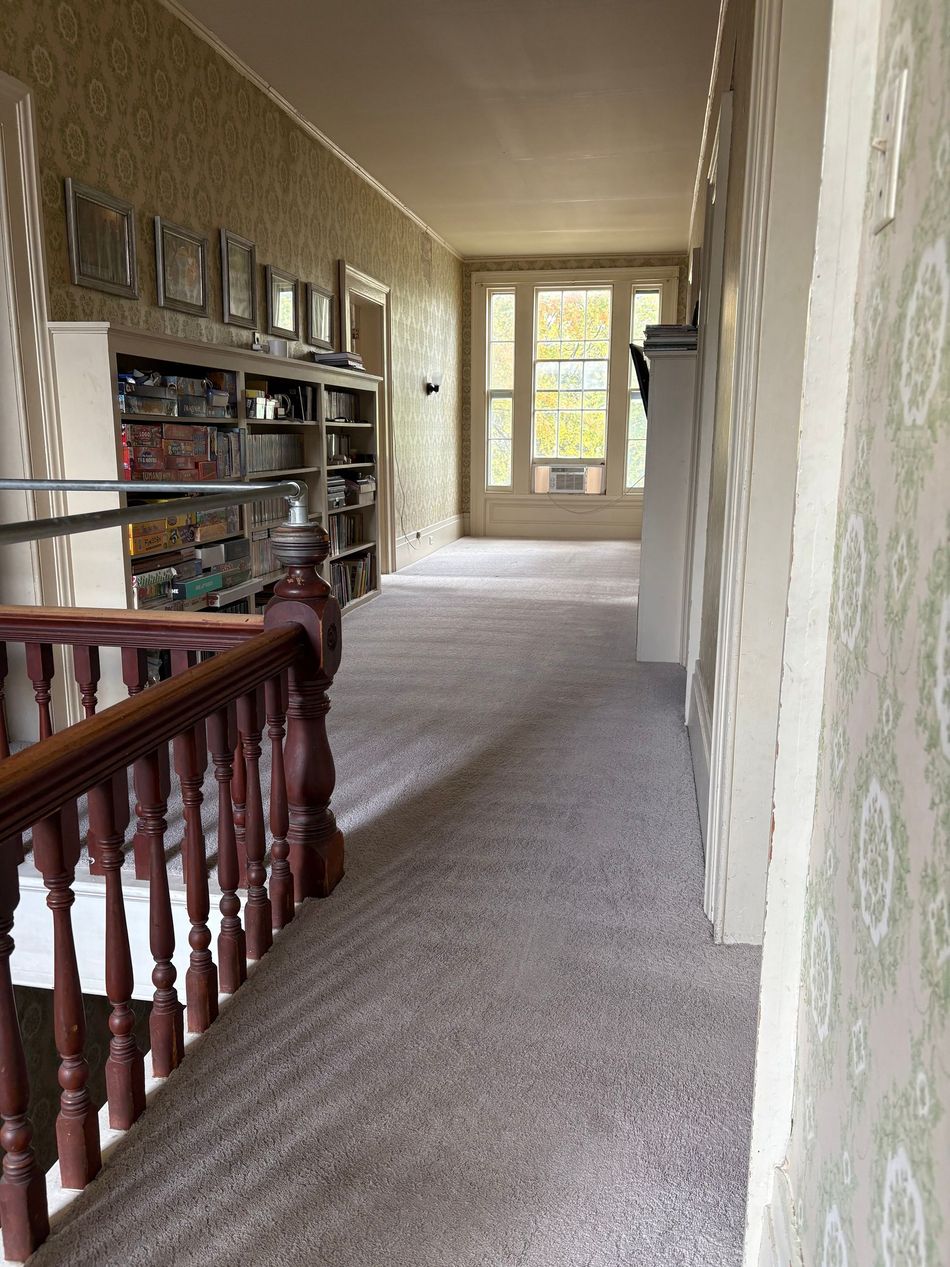Hallway with bookshelves, staircase, and a window at the end. Carpeted floor, light-colored walls, and a wooden banister.