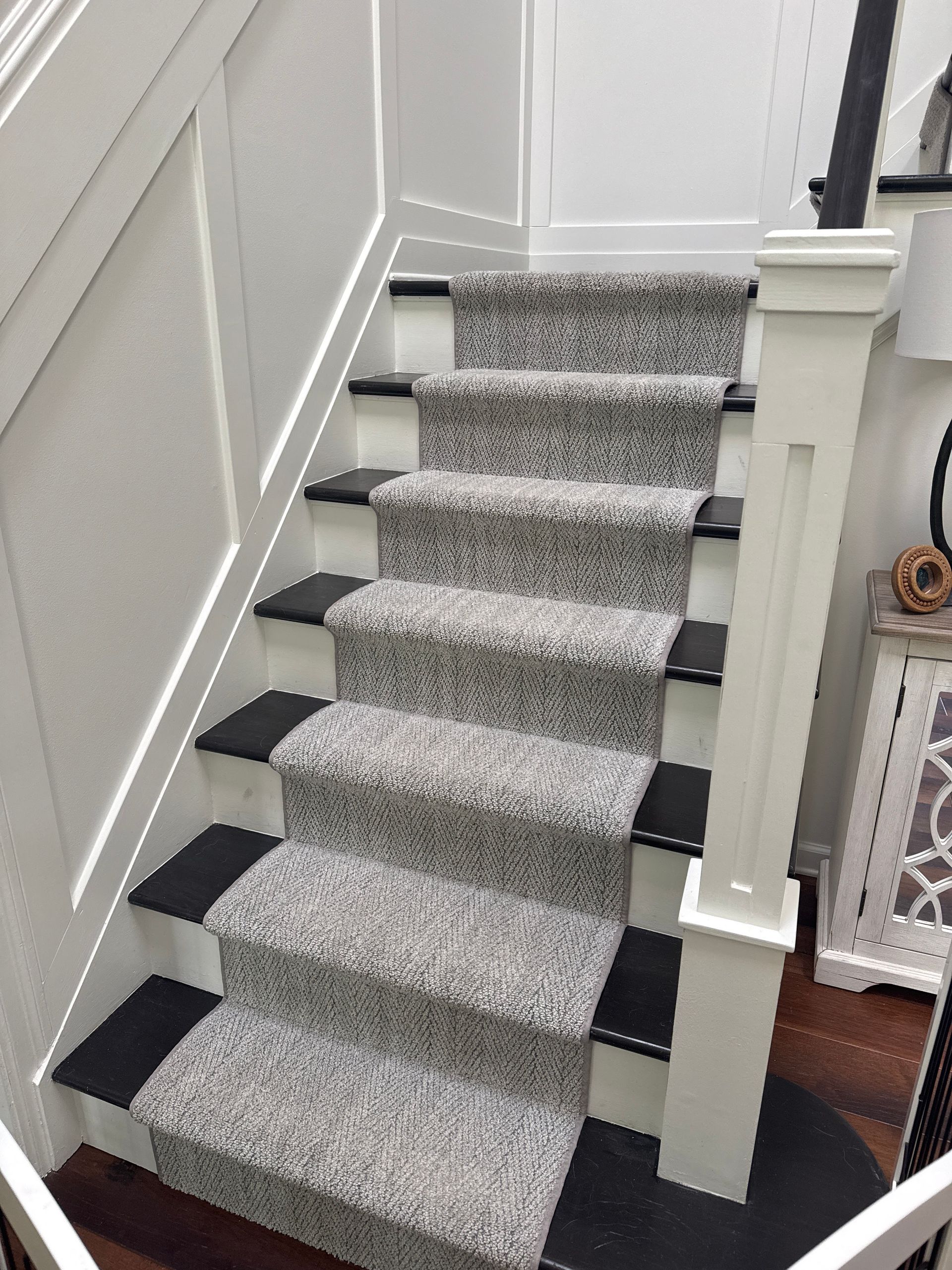 Staircase with gray carpet runner on black treads, white walls and trim, hardwood floor.