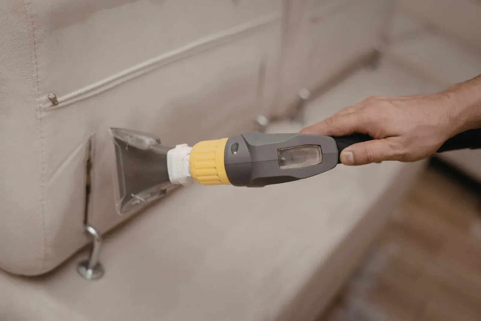 A person cleaning a light-colored sofa with a handheld upholstery cleaner.